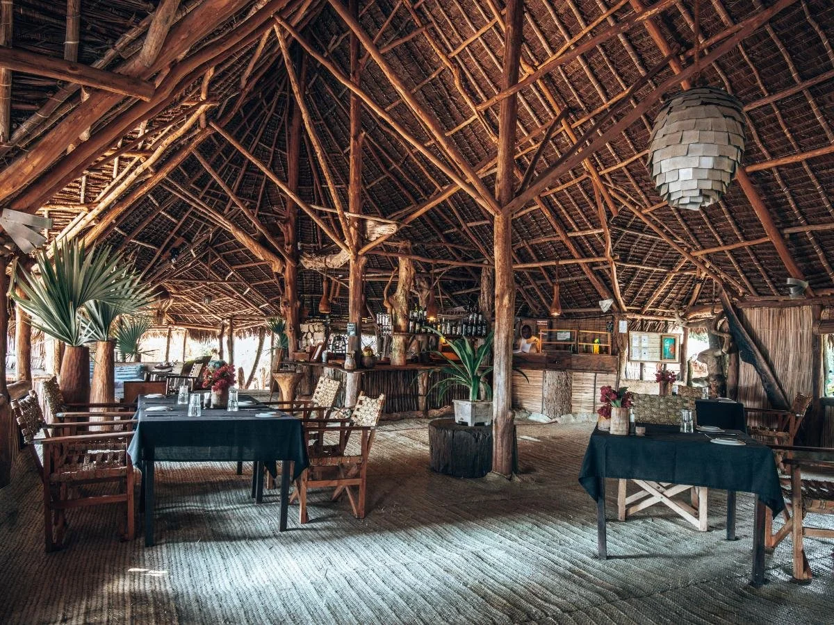 Ristorante rustico all'aperto con travi in ​​legno e tetto di paglia. I tavoli sono apparecchiati con tovaglie nere e circondati da sedie intrecciate, creando un'atmosfera accogliente e naturale.