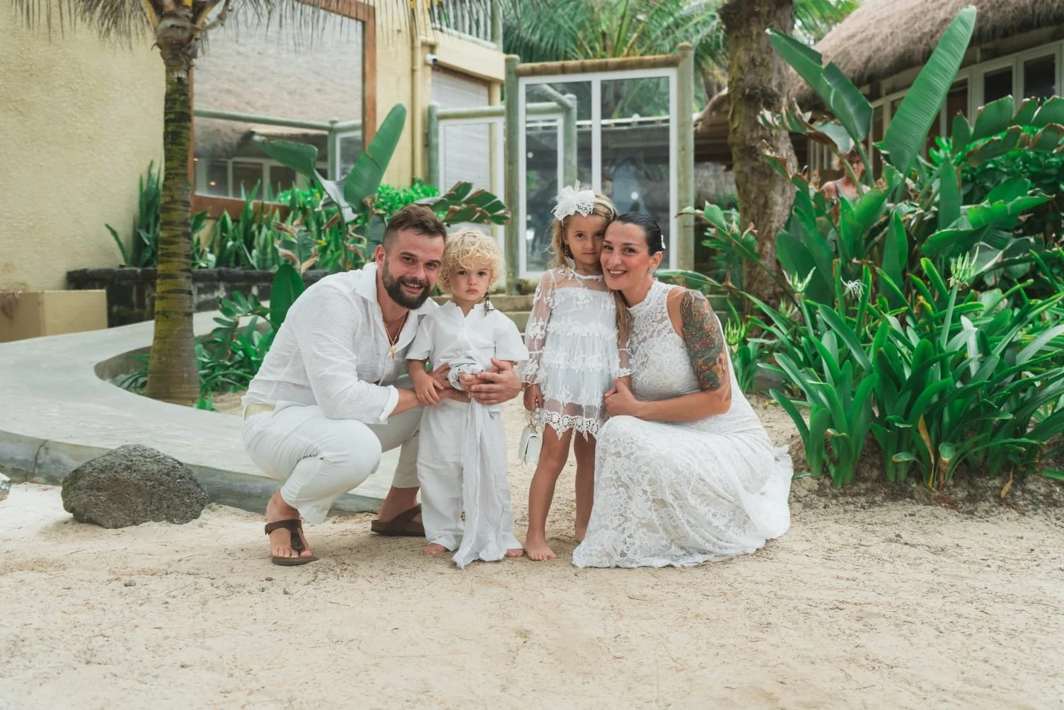 Sposarsi alle Mauritius. Una famiglia di quattro persone vestita di bianco posa su un sentiero sabbioso circondato da rigogliose piante tropicali. L'atmosfera è tranquilla e gioiosa, e suggerisce una festa.