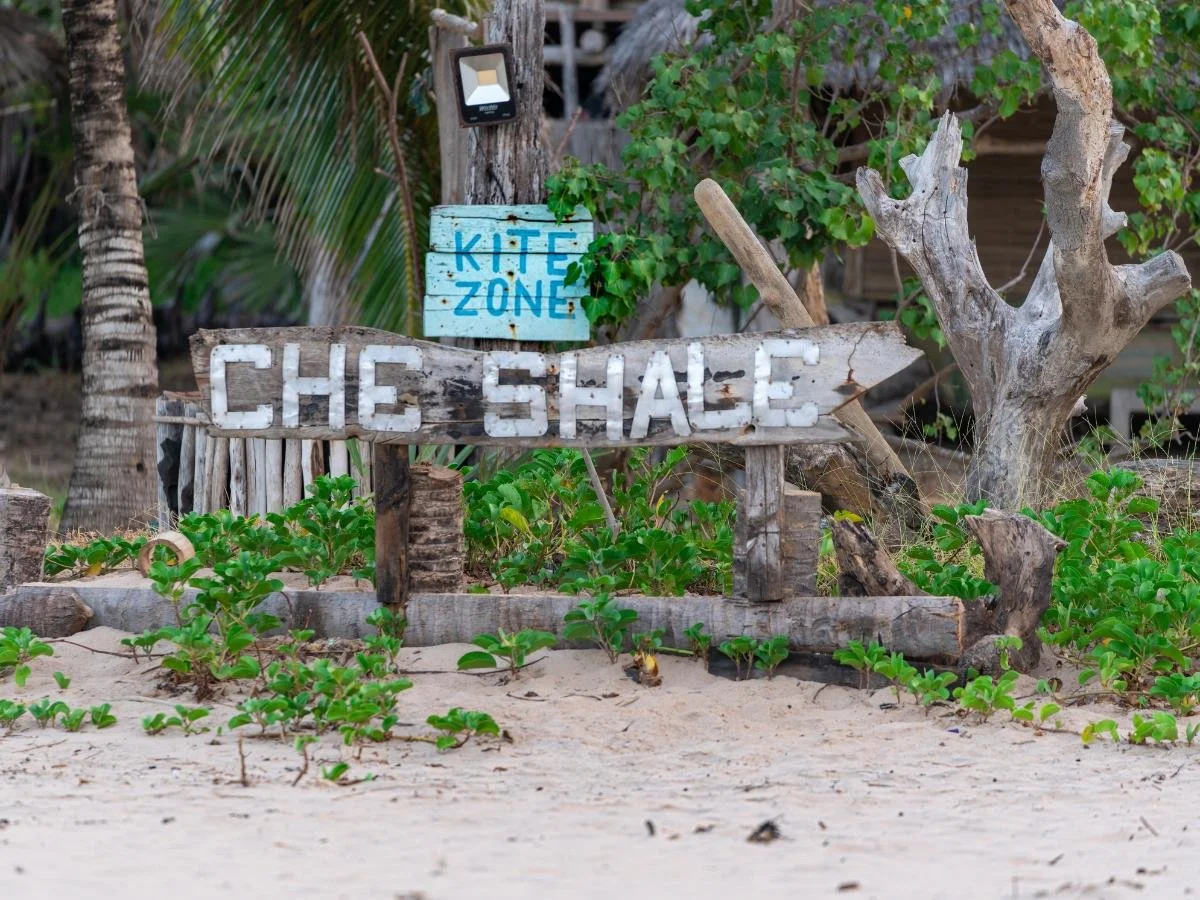 Un cartello in legno invecchiato su una spiaggia sabbiosa recita "Che Shale" e "Kite Zone", circondato da una lussureggiante vegetazione verde e palme, evocando un'atmosfera tropicale. È il luogo perfetto dove fare kitesurf in kenya.