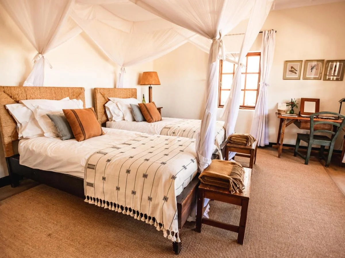 Interno di una luminosa camera da letto in stile safari. Ci sono due letti singoli con zanzariere bianche, testiere in vimini e coperte decorate con motivi geometrici.