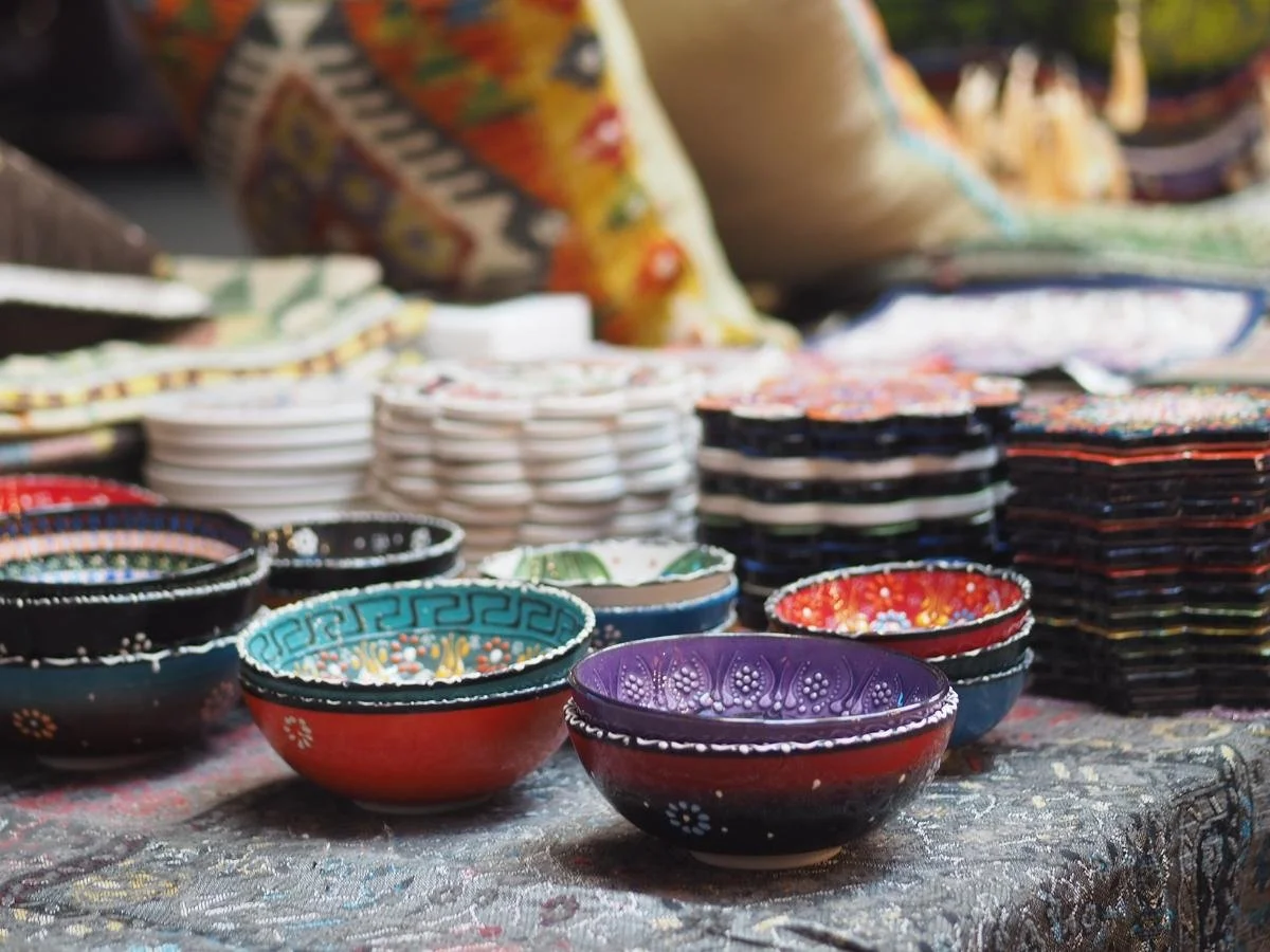 Ciotole di ceramica colorate con motivi intricati sono appoggiate su una tovaglia decorata. Pile di piatti e tessuti dai colori vivaci creano un'atmosfera calda e artigianale.