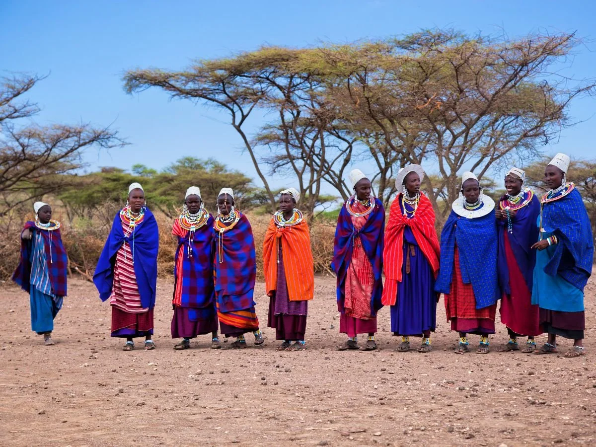 Un gruppo di persone in vivaci abiti tradizionali è in piedi su un sentiero sterrato, con abiti colorati e accessori di perline. Alberi di acacia e un cielo azzurro e terso fanno da sfondo.