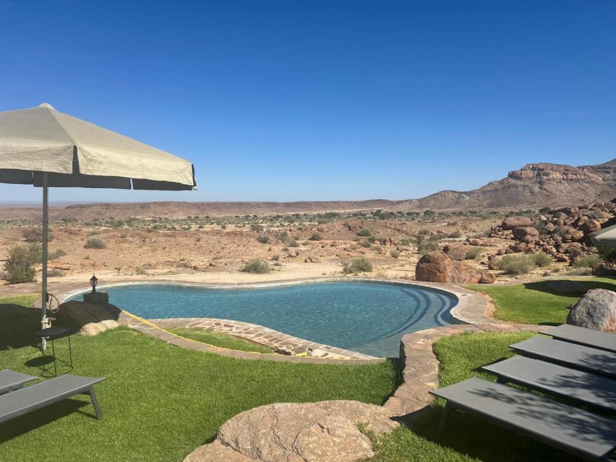 Una piscina turchese a forma di cuore con bordi in pietra, situata in un lussureggiante giardino verde con sedie a sdraio e un ombrellone beige, che si affaccia su un vasto paesaggio desertico montuoso e roccioso sotto un cielo blu limpido.
