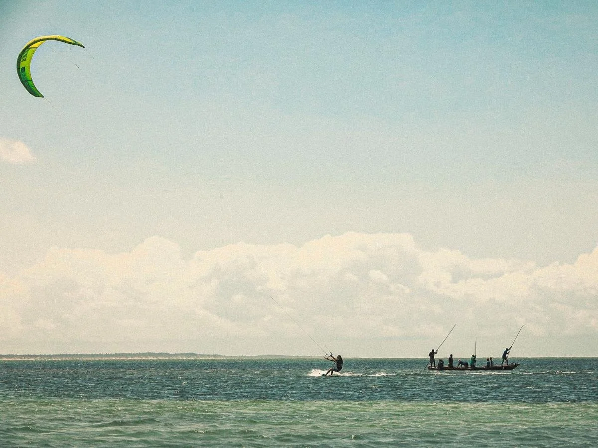 Un kitesurfer scivola sull'oceano vicino a una barca da pesca con diverse persone che tengono in mano delle canne. La scena è serena sotto un cielo azzurro con nuvole sparse.