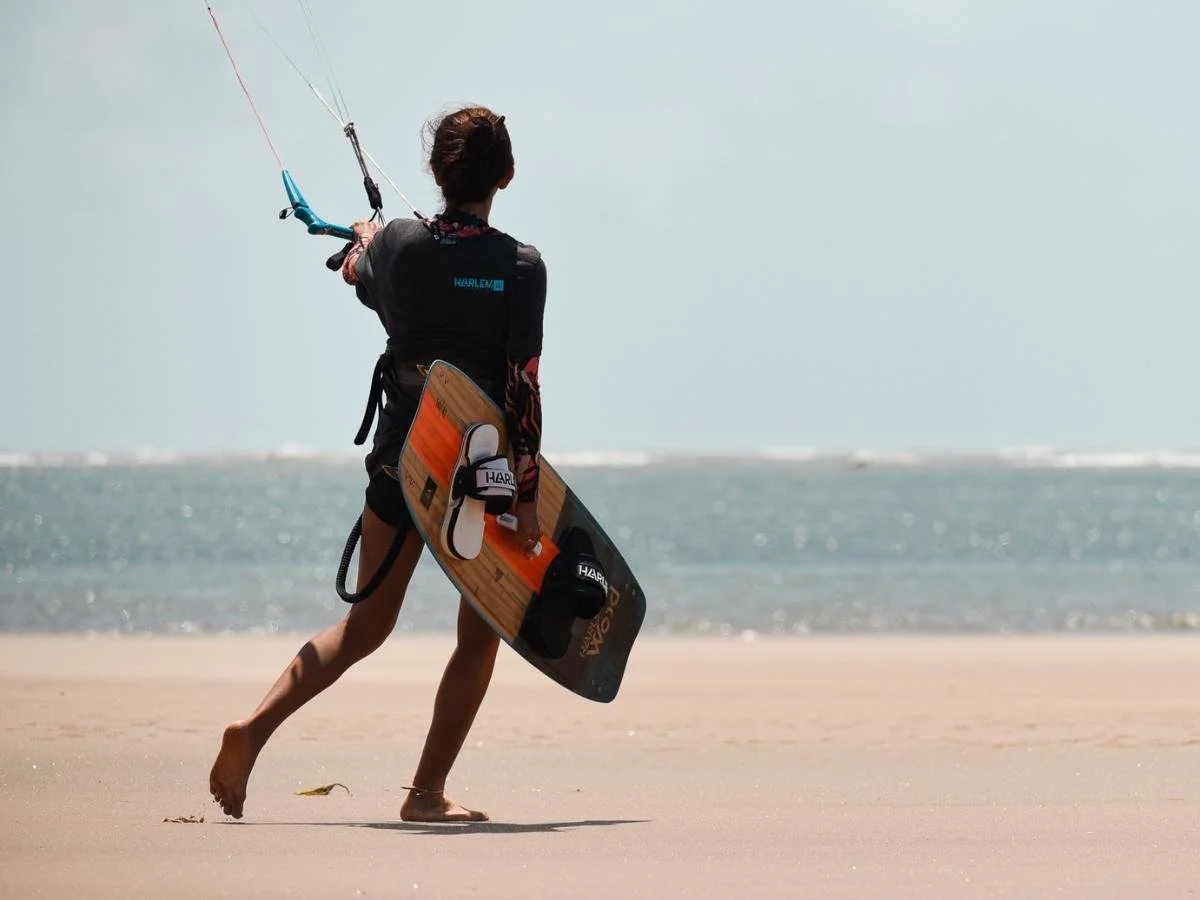 Una persona cammina a piedi nudi su una spiaggia sabbiosa, con in mano una tavola da kitesurf e i fili dell'aquilone, di fronte all'oceano. La scena trasmette avventura e libertà.