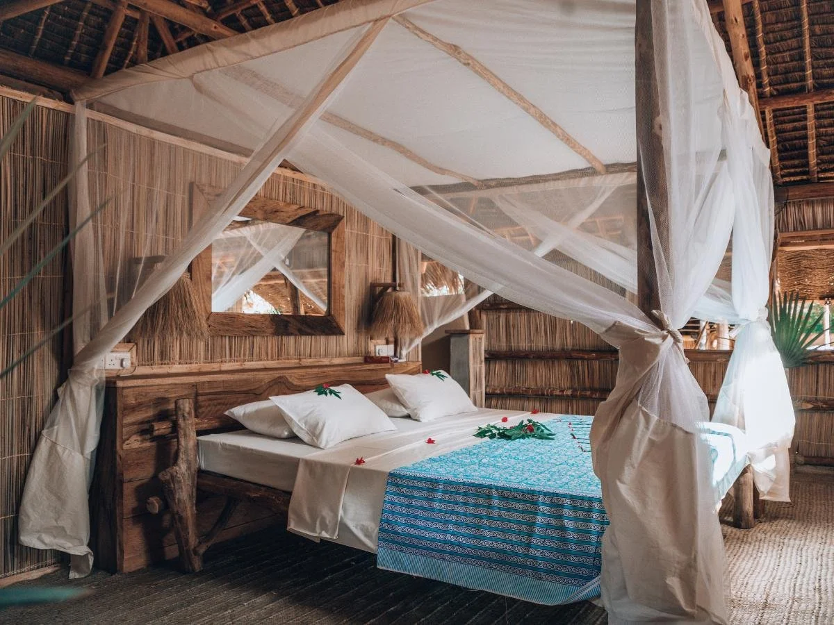 Camera da letto rustica con letto in legno, baldacchino bianco e copriletto con fantasia blu. Arredamento naturale, toni caldi, atmosfera accogliente e serena. È il luogo perfetto per alloggiare dopo una sessione di kitesurf in Kenya.