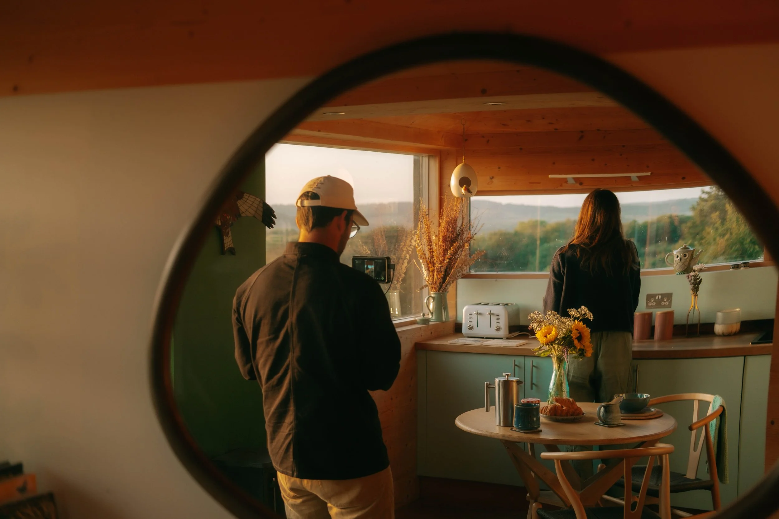 A man and a woman are in a kitchen, viewed through a circular window. The man is standing and recording with a camera, wearing glasses and a cap. The woman is facing the window, looking outside at a scenic landscape with trees. The kitchen has a roun