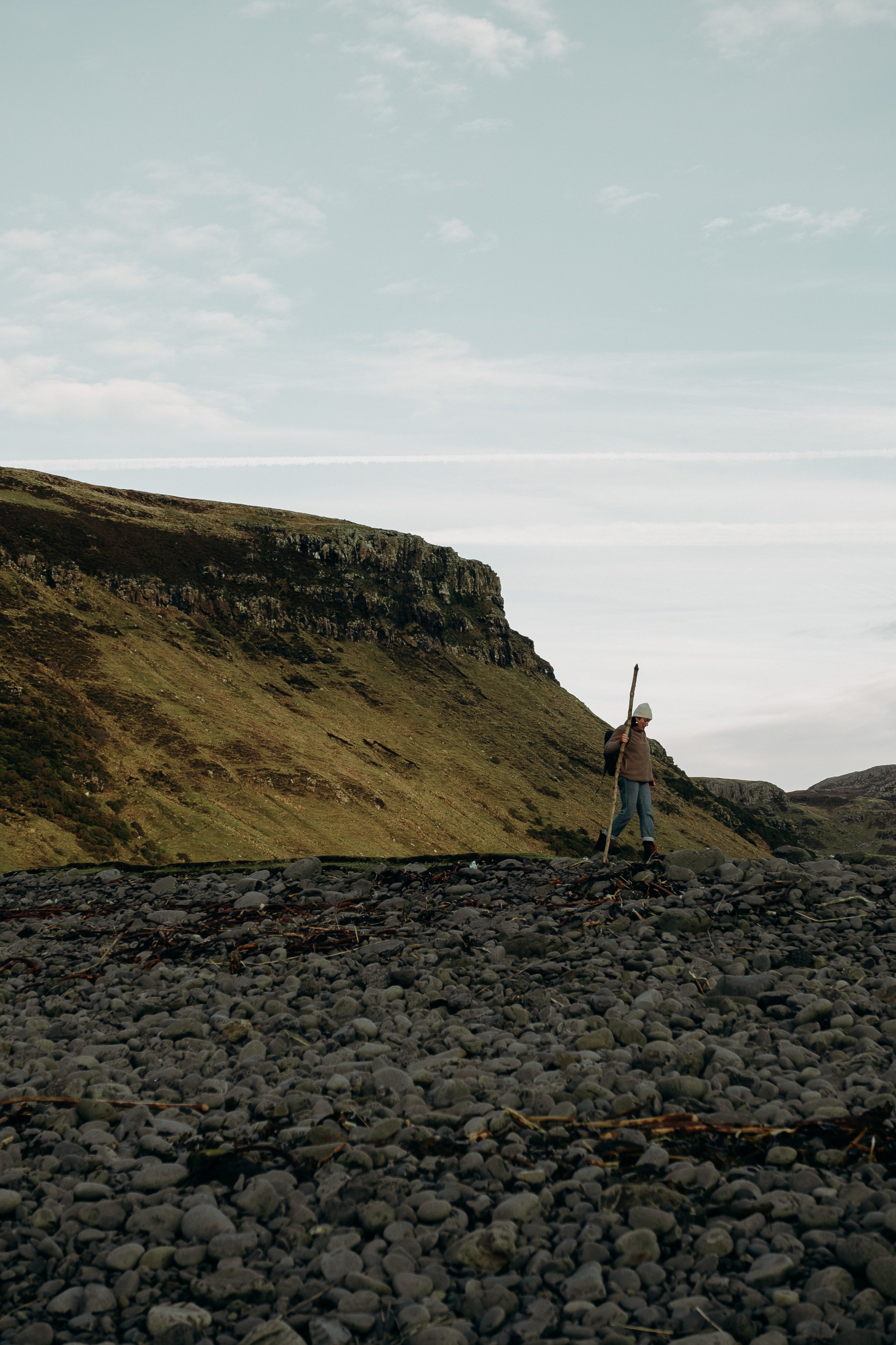 A person wearing a gray hat, brown jacket, and blue jeans walking on a rocky beach with a tall stick, mountains in the background, and a cloudy sky.