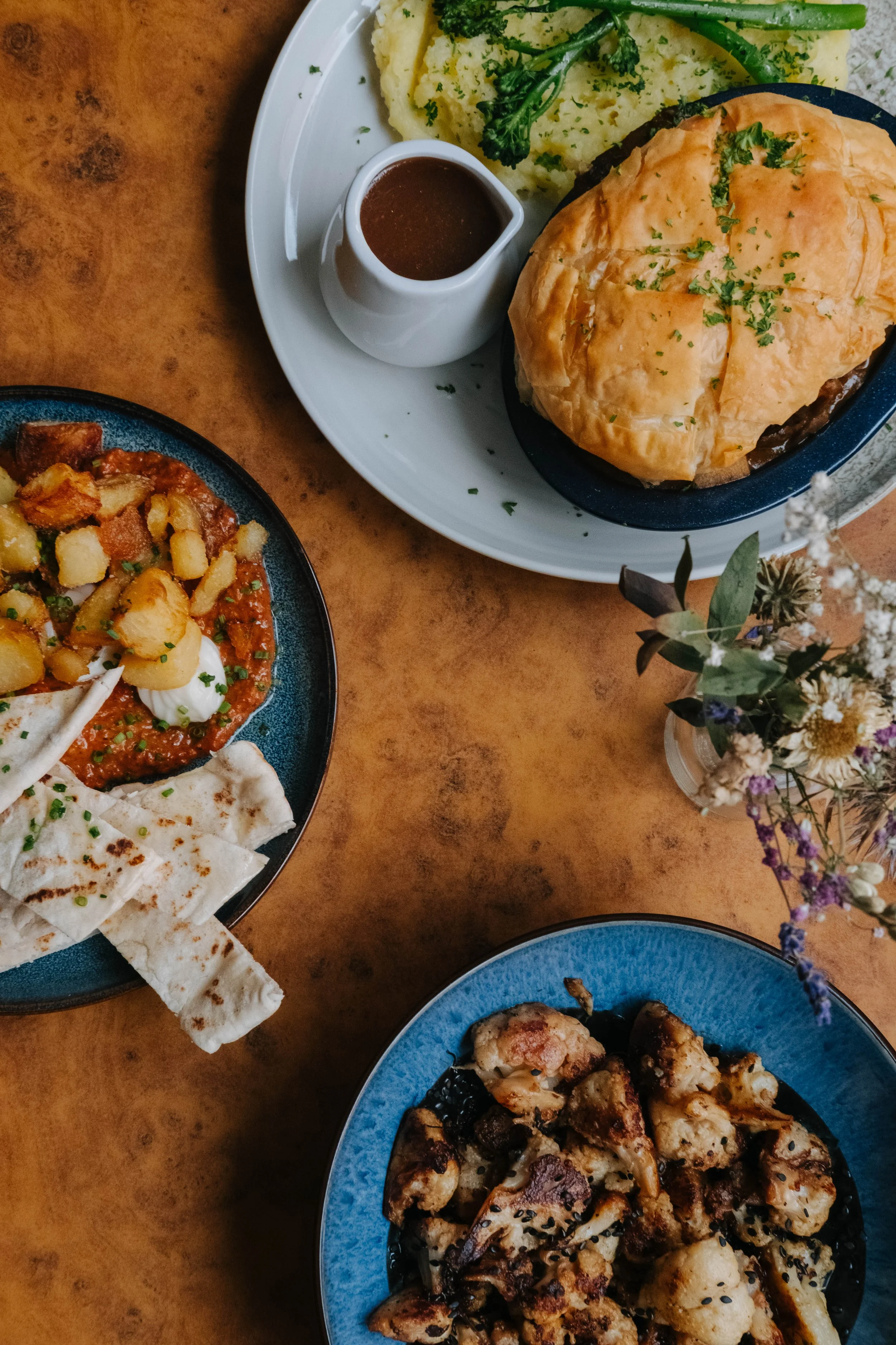 A top view of a table with three dishes: a plate of grilled chicken with black sesame seeds, a bowl of roasted cauliflower, and a plate of Mexican food including chilaquiles with green sauce, fried eggs, potatoes, pico de gallo, and flour tortillas.