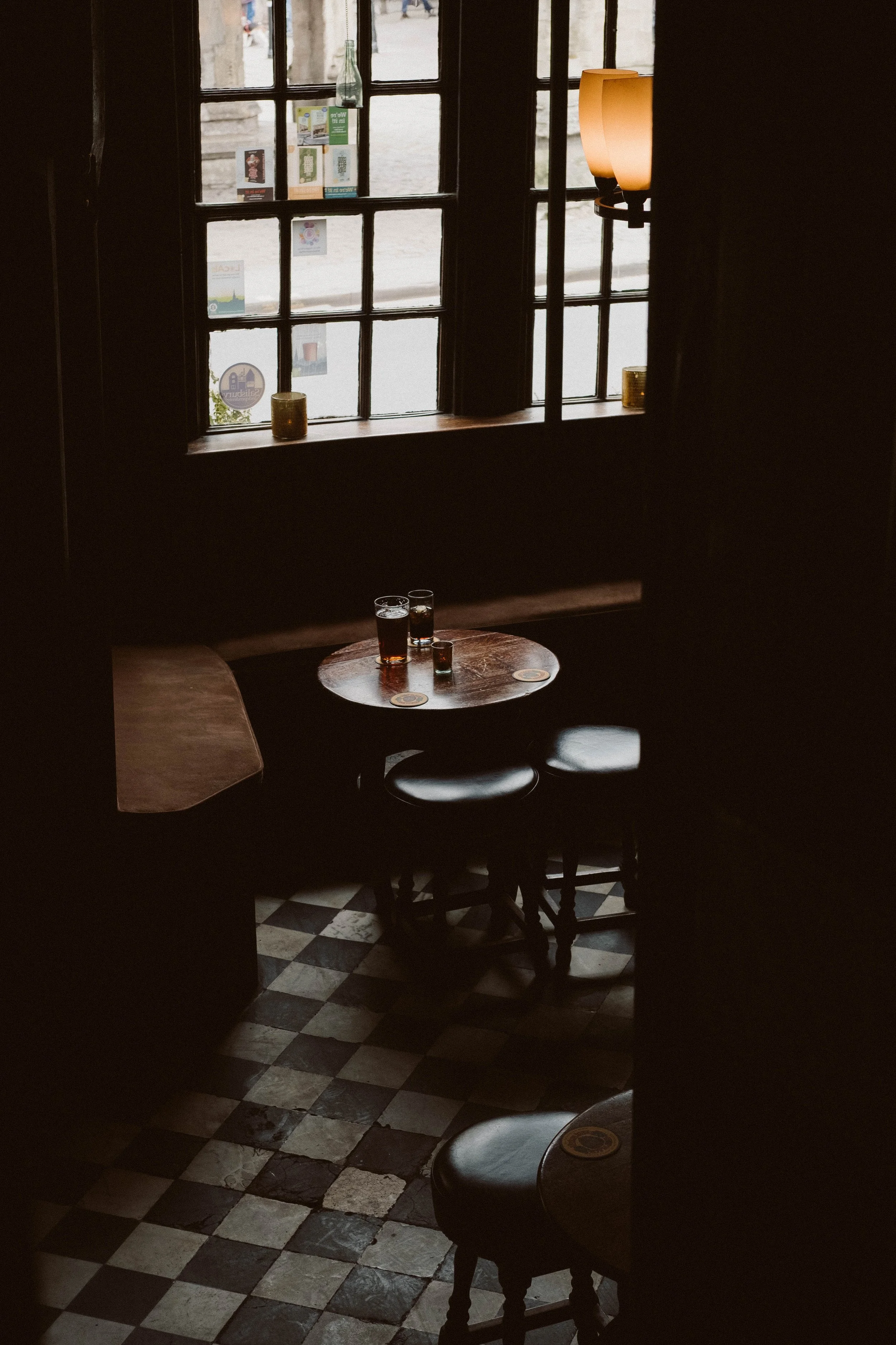 A cozy bar or pub interior with a small round table near a window. The table has three glasses of dark beer and a small shot glass. Sunlight streams through the window, illuminating the dark wood furniture and black and white checkered floor tiles. T