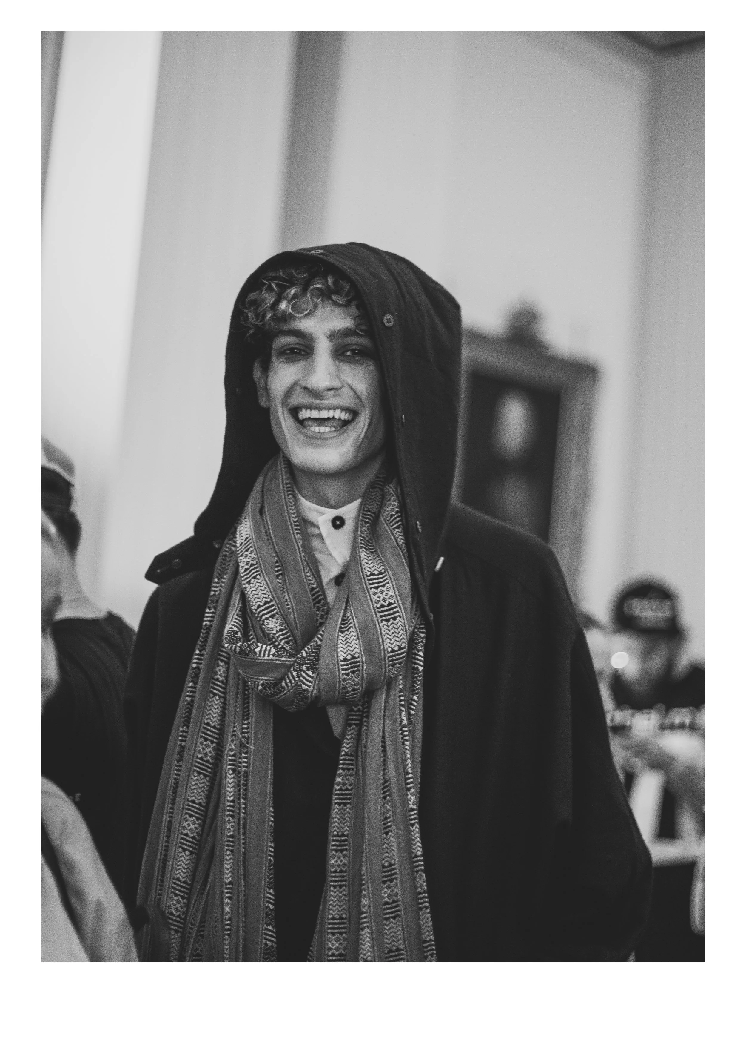 A person with curly hair smiling and wearing a hooded jacket and patterned scarf, indoors with blurred background.