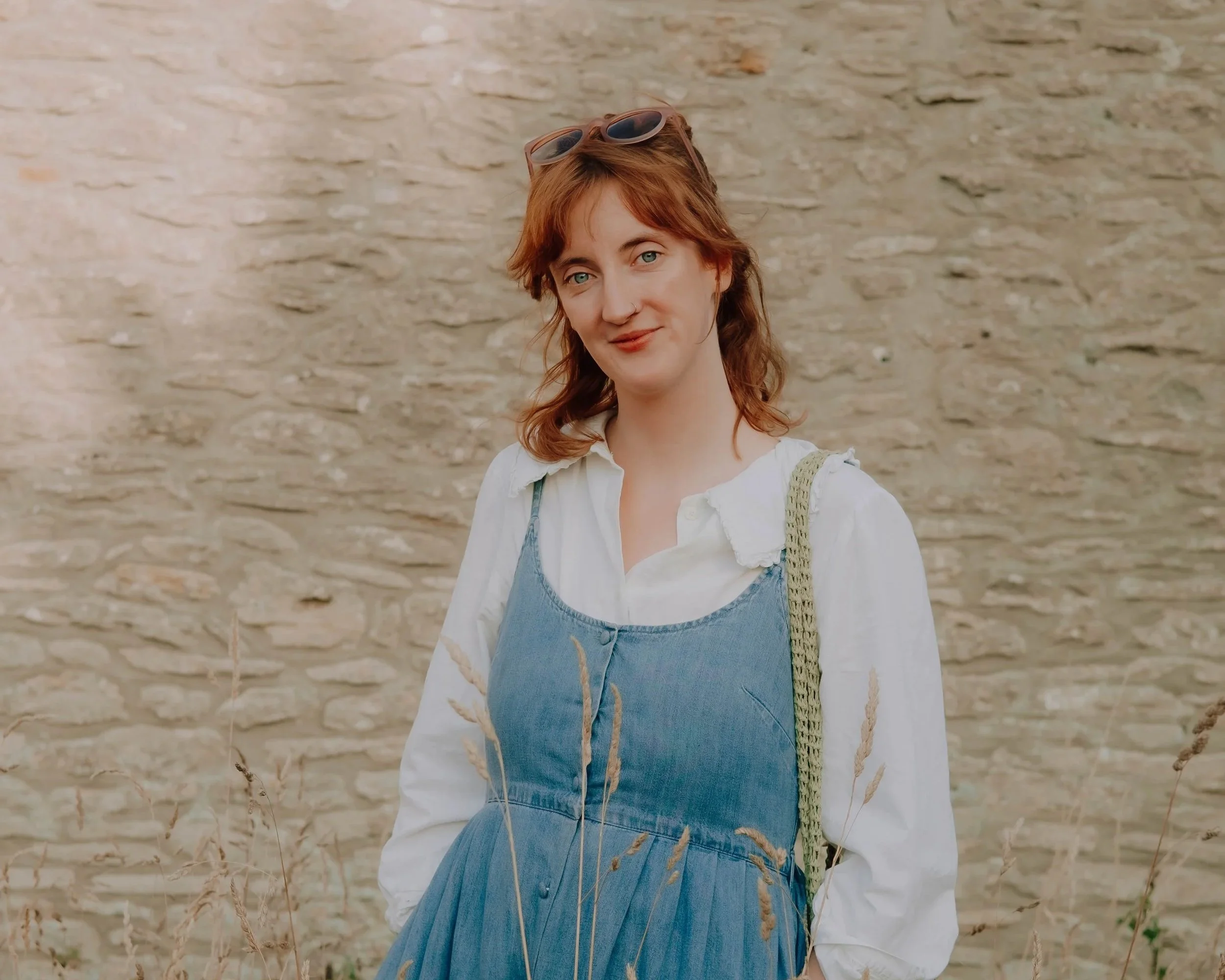 A woman with curly red hair and blue eyes, wearing a white blouse, denim overall dress, and sunglasses on her head, standing outdoors in front of a stone wall.