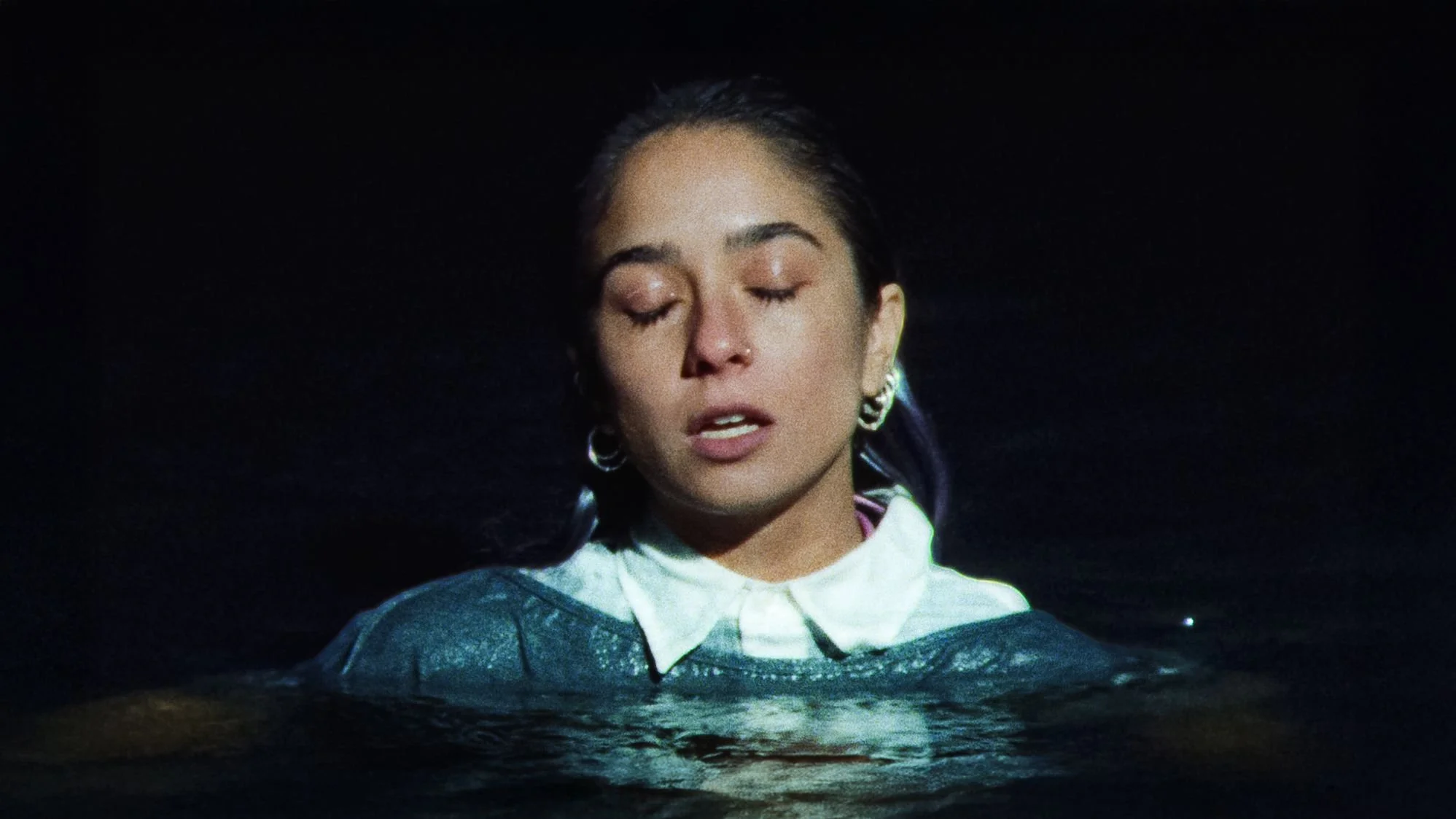 A woman with dark hair and earrings, wearing a collared shirt and sweater, stands in water with her eyes closed, against a dark background.
