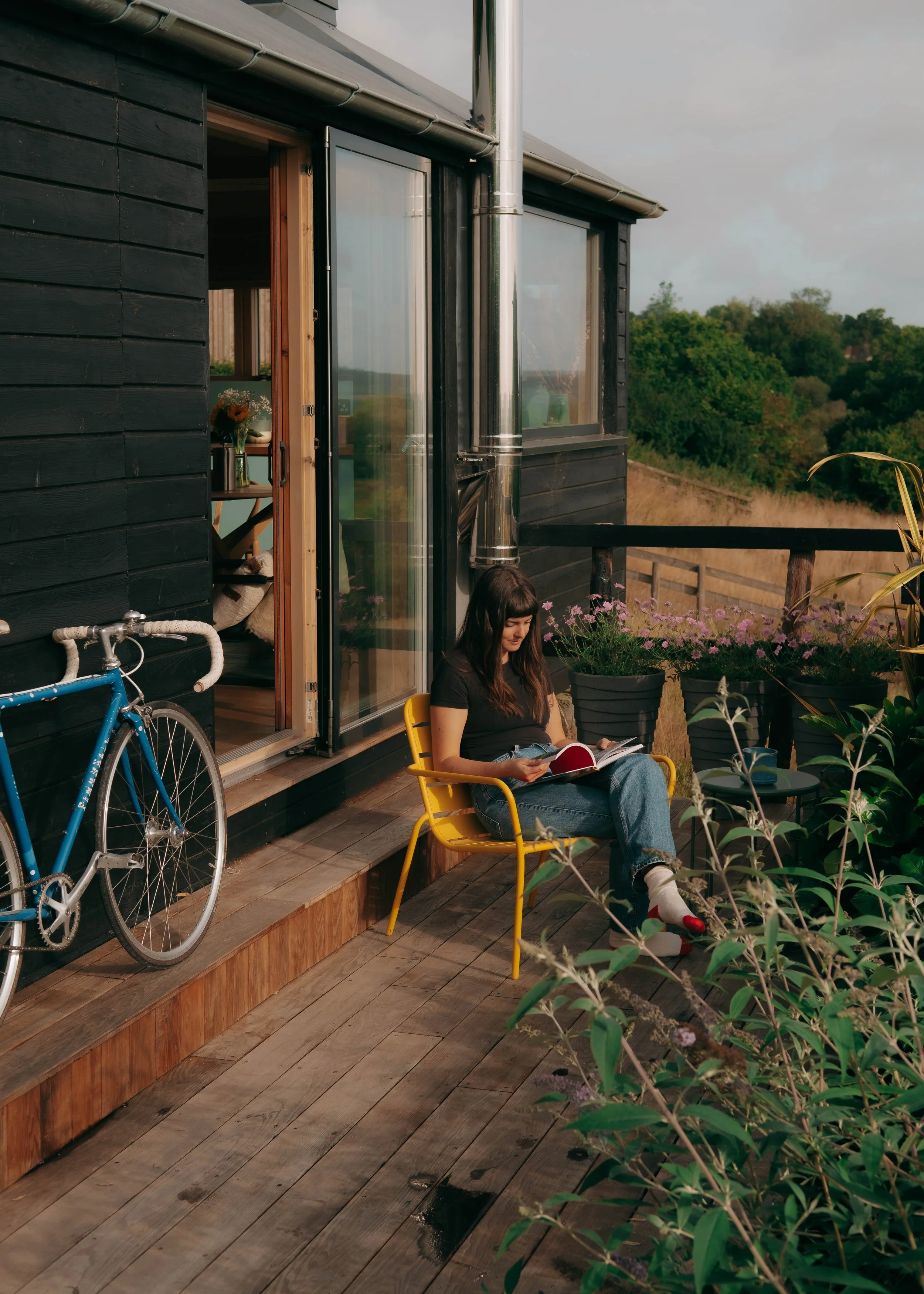 A woman with long dark hair sitting in a yellow chair on a wooden deck, reading a book. She is wearing jeans, a black t-shirt, and colorful socks. To her left, there's a blue bicycle leaning against the black exterior wall of a house. The house has a