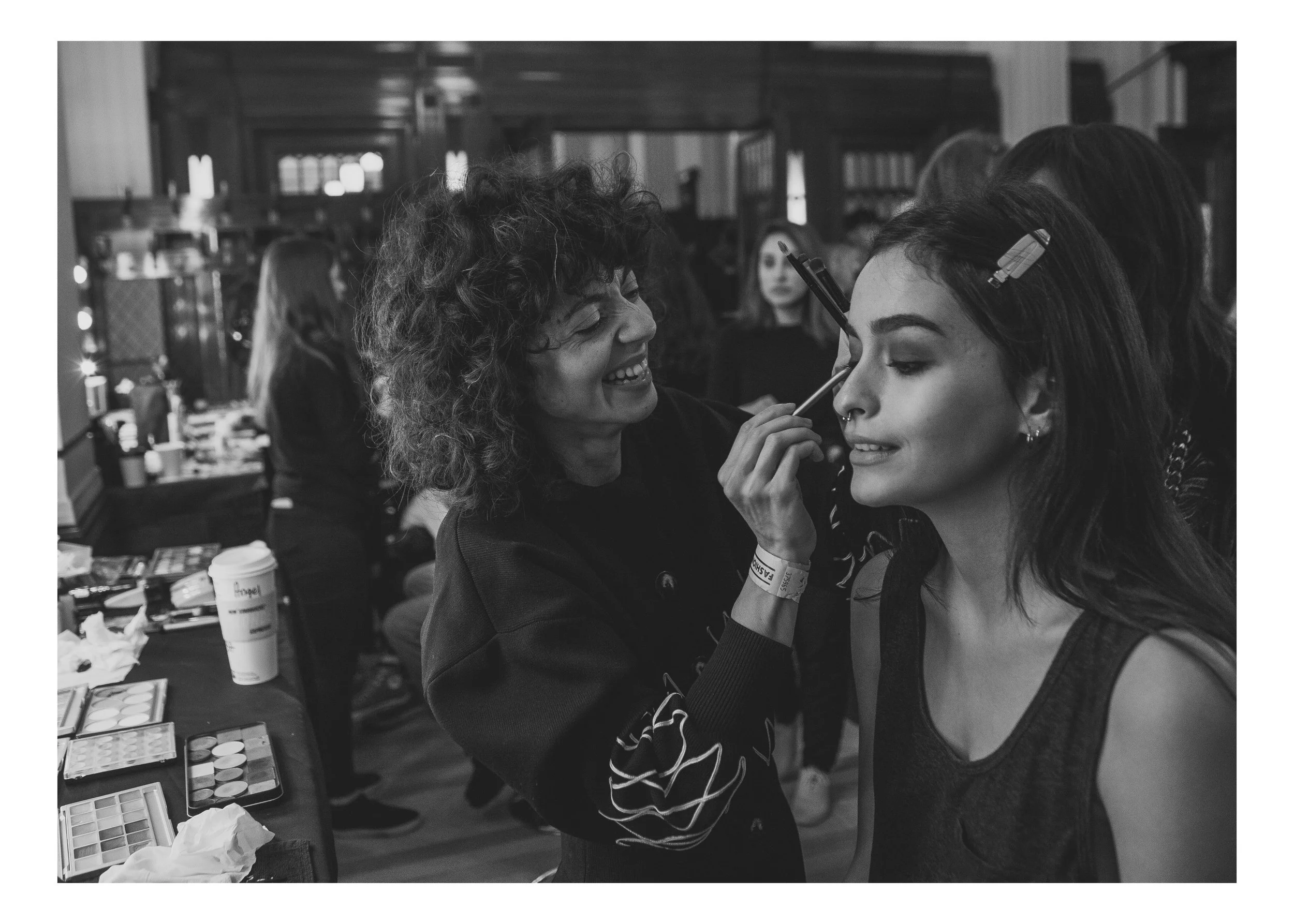 A makeup artist with curly hair applies eyeshadow to a woman with long dark hair, who is wearing a sleeveless top, in a crowded indoor setting.