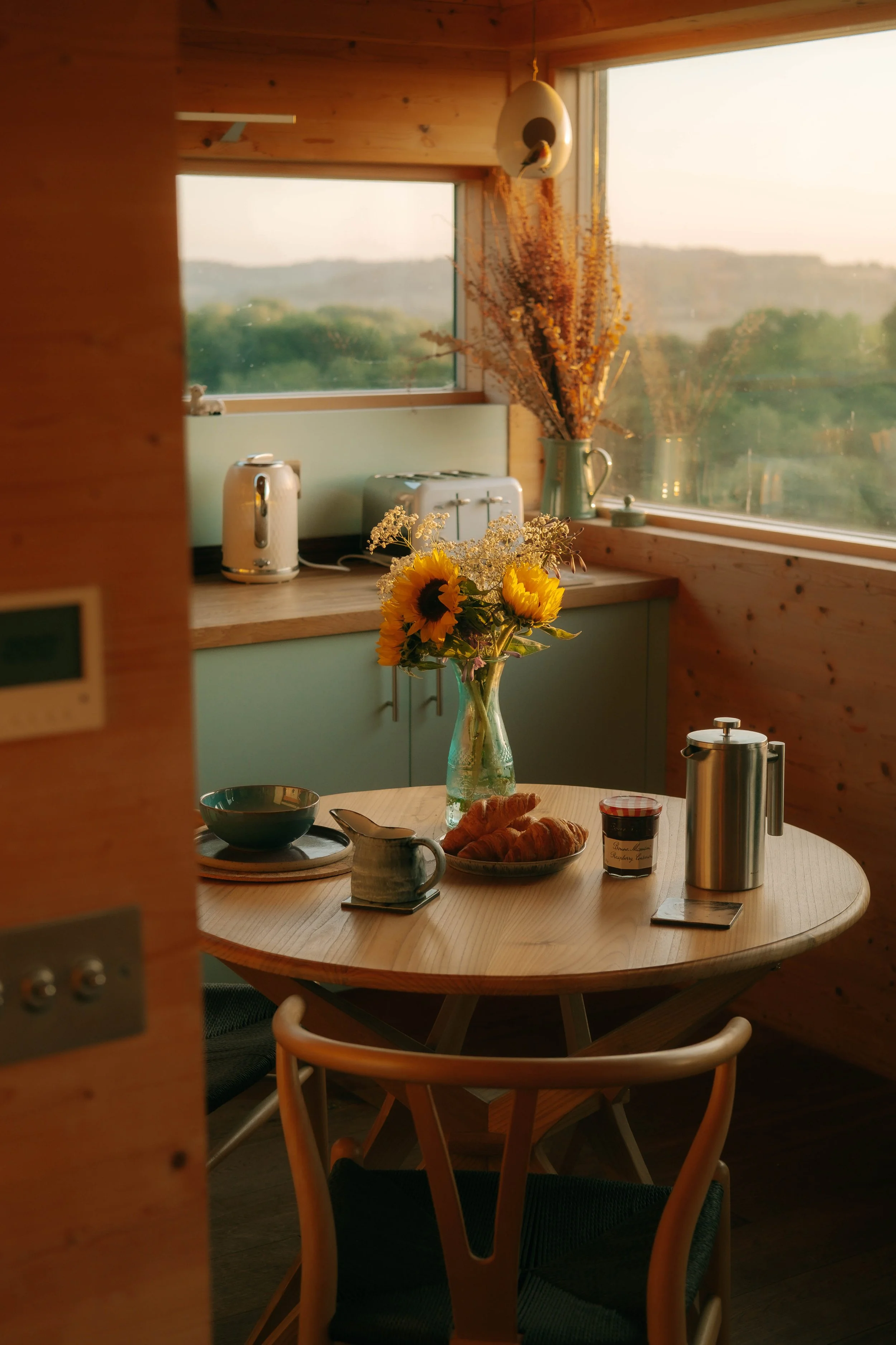 A cozy breakfast nook with a round wooden table, a vase of sunflowers, croissants, a cup of jam, a stainless steel teapot, and a ceramic mug, illuminated by natural sunlight through large windows showing a scenic landscape.