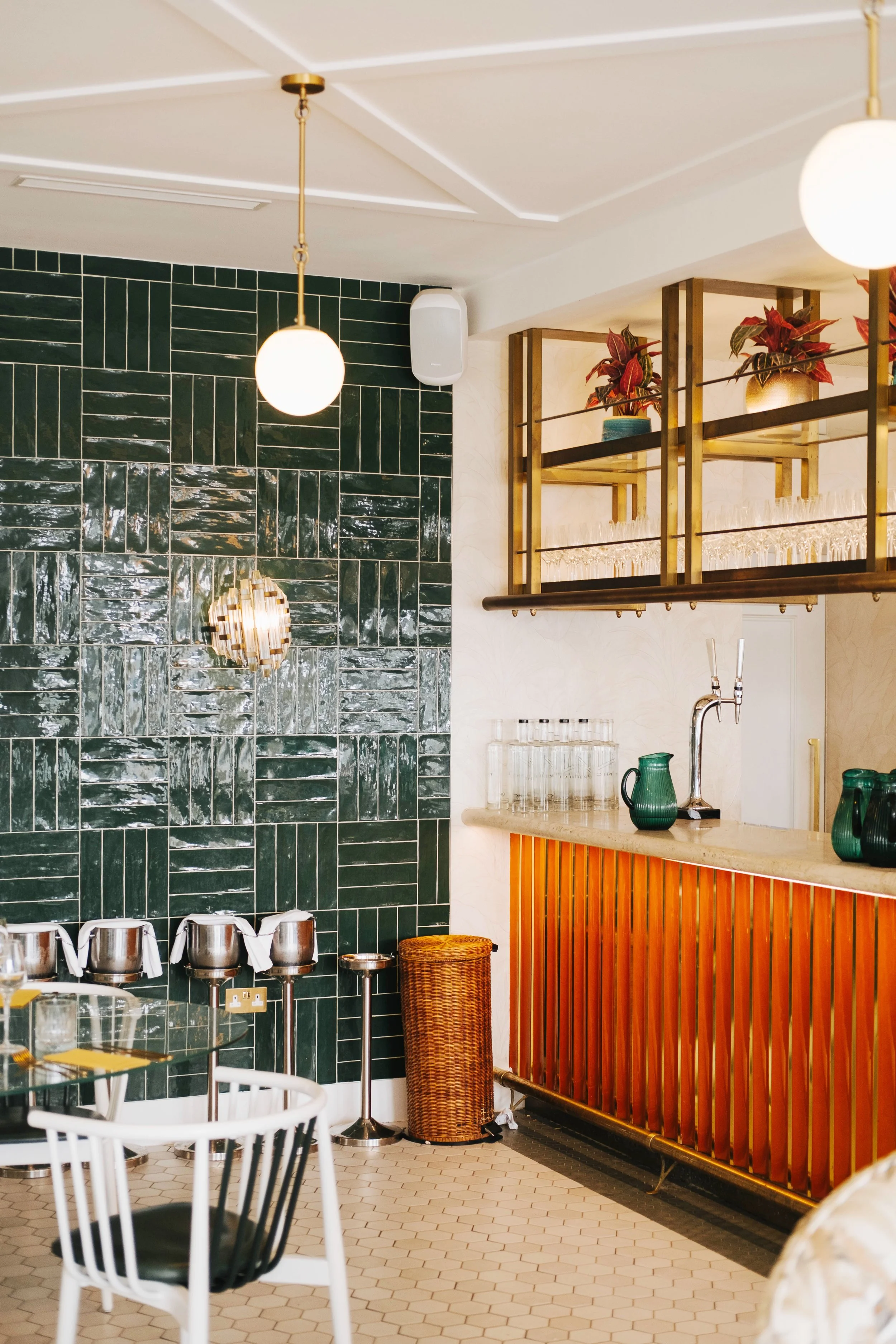 Interior of a stylish bar or restaurant with dark green tiled wall, hanging lights, wooden bar counter with green pitchers, glassware, and decorative plants on shelves.