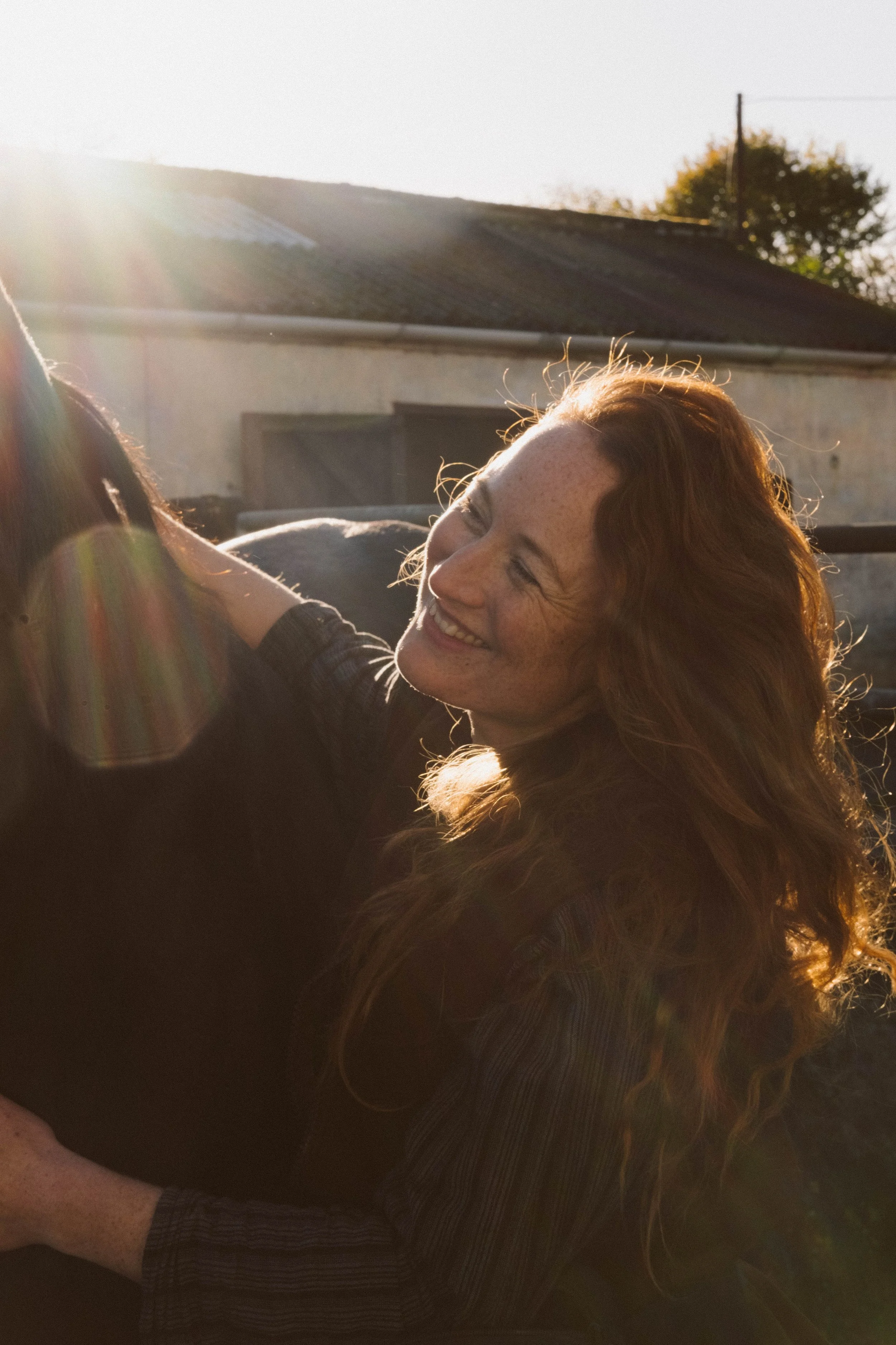 A woman with red hair smiling and hugging a dark-colored horse outside during sunset.