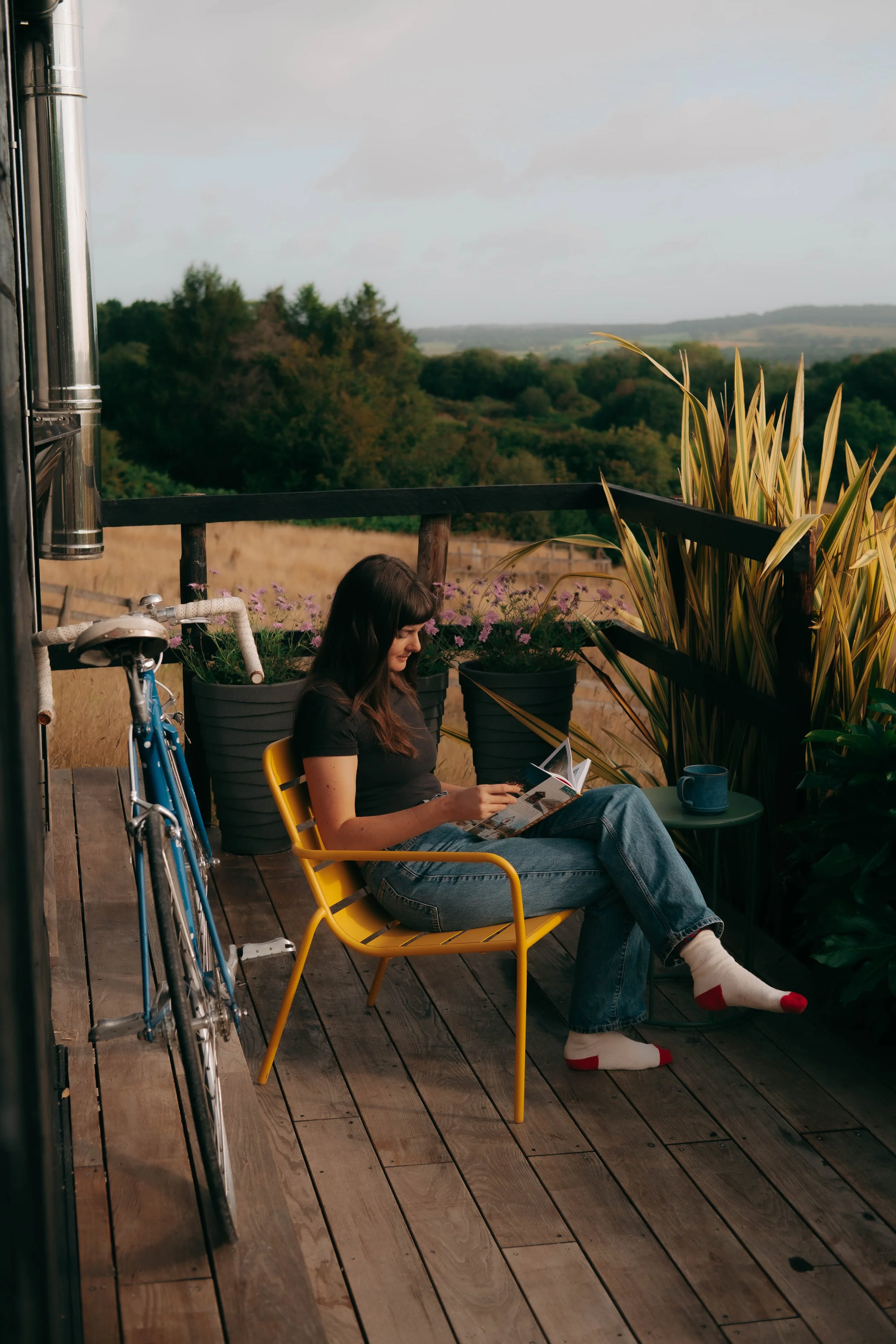 A woman sitting on a yellow chair on a wooden deck, reading a magazine with a scenic countryside view in the background, and a bicycle leaning against the side of the deck.