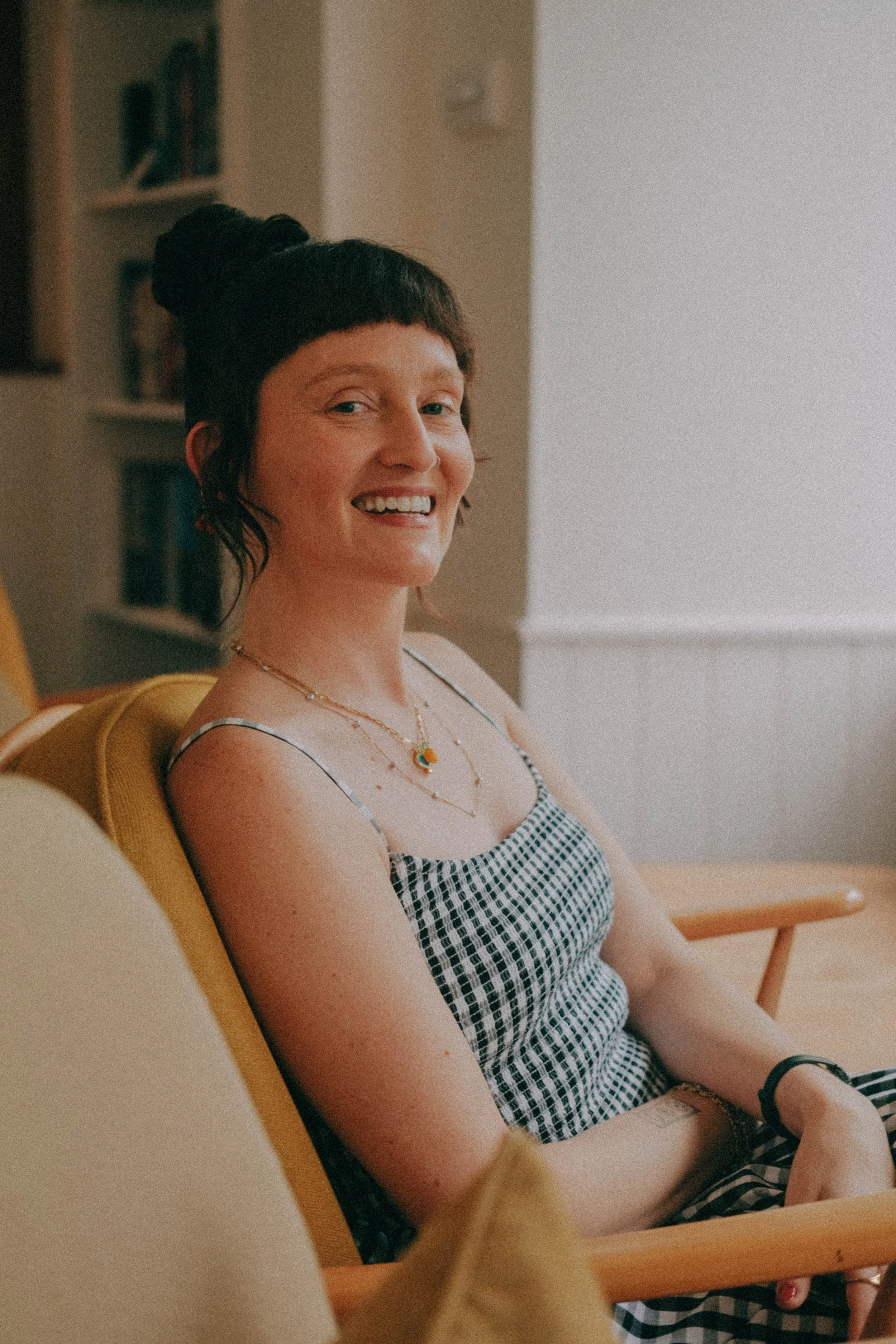 A woman with short dark hair in a bun, smiling while sitting on a yellow armchair in a cozy room. She is wearing a checkered tank top and layered necklaces.