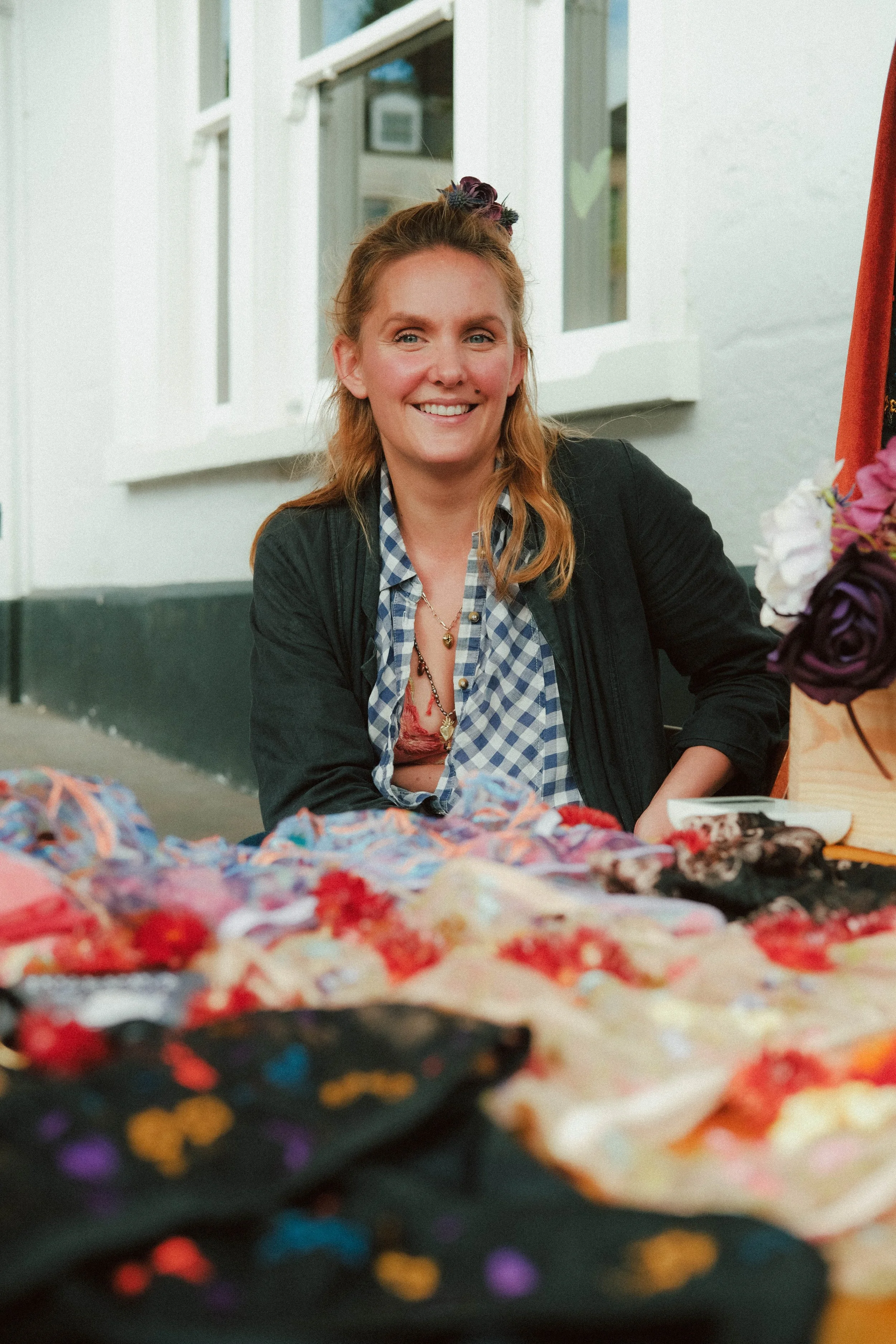 A smiling woman with reddish hair and a floral headband sitting at a table filled with colorful fabrics and flowers.