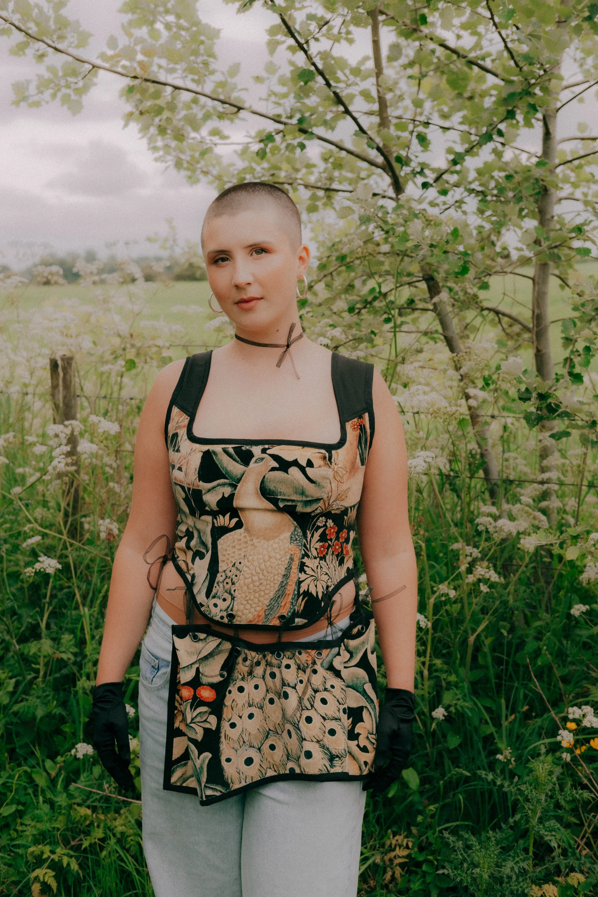 A young woman with a shaved head, wearing a black choker, hoop earrings, black gloves, and a dress with a peacock print, standing outdoors with lush green plants and trees in the background.