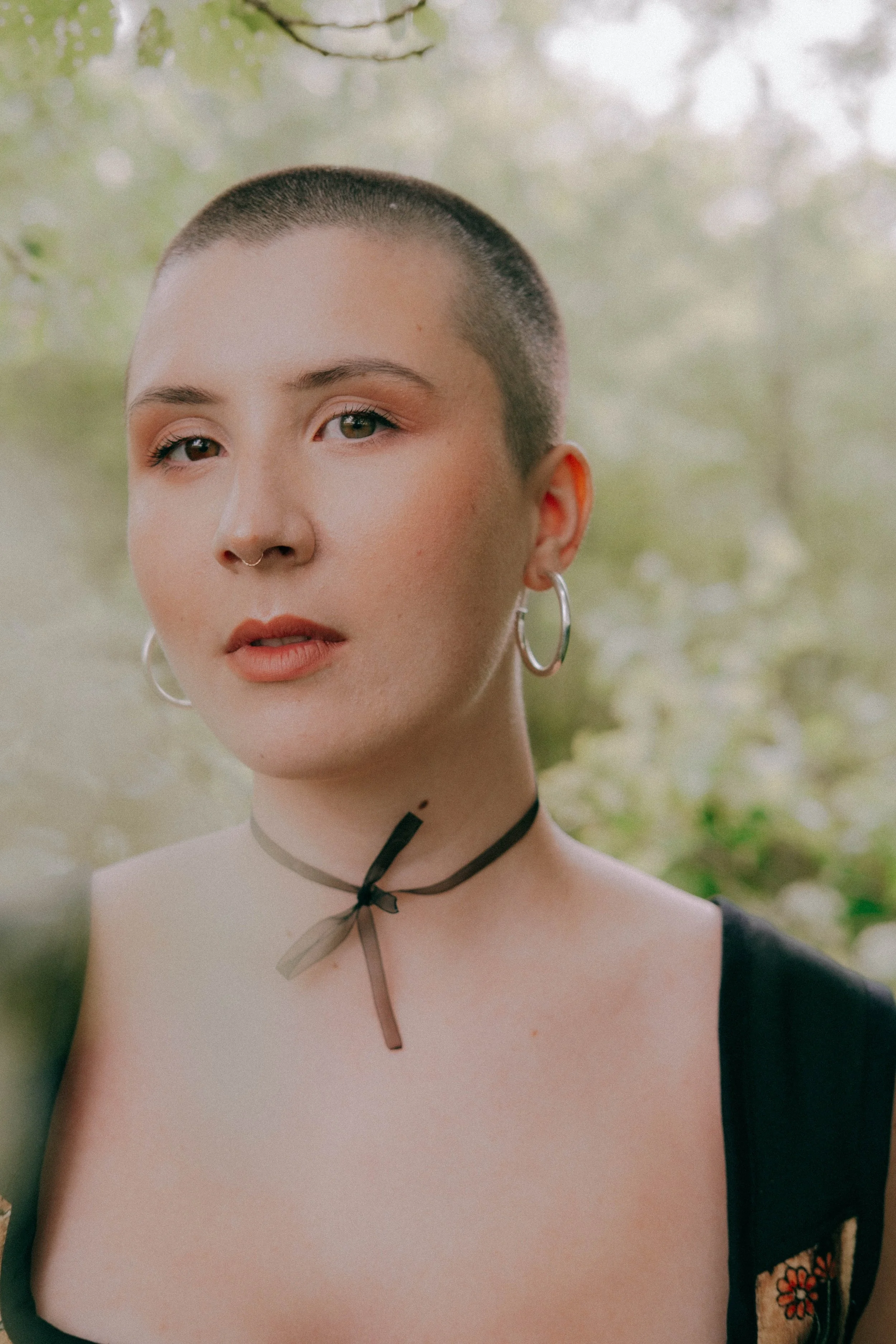 Portrait of a woman with short buzzed hair, wearing hoop earrings, a septum piercing, a black choker with a bow, and a black top with floral embroidery, outdoors with blurred green foliage in the background.