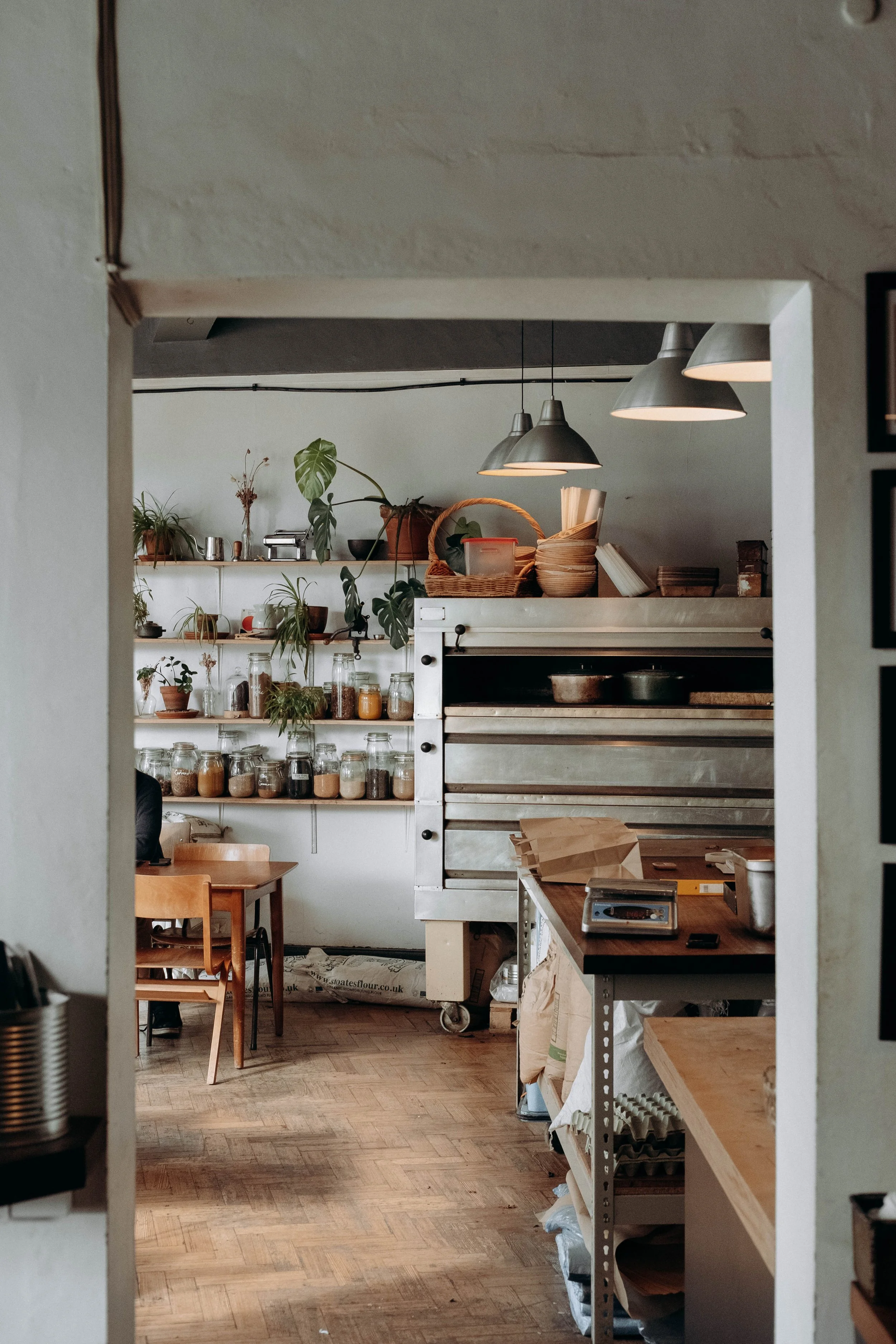 Interior view of a cozy kitchen or bakery with wooden furniture, shelves with jars and plants, and metallic kitchen equipment, seen through a doorframe.