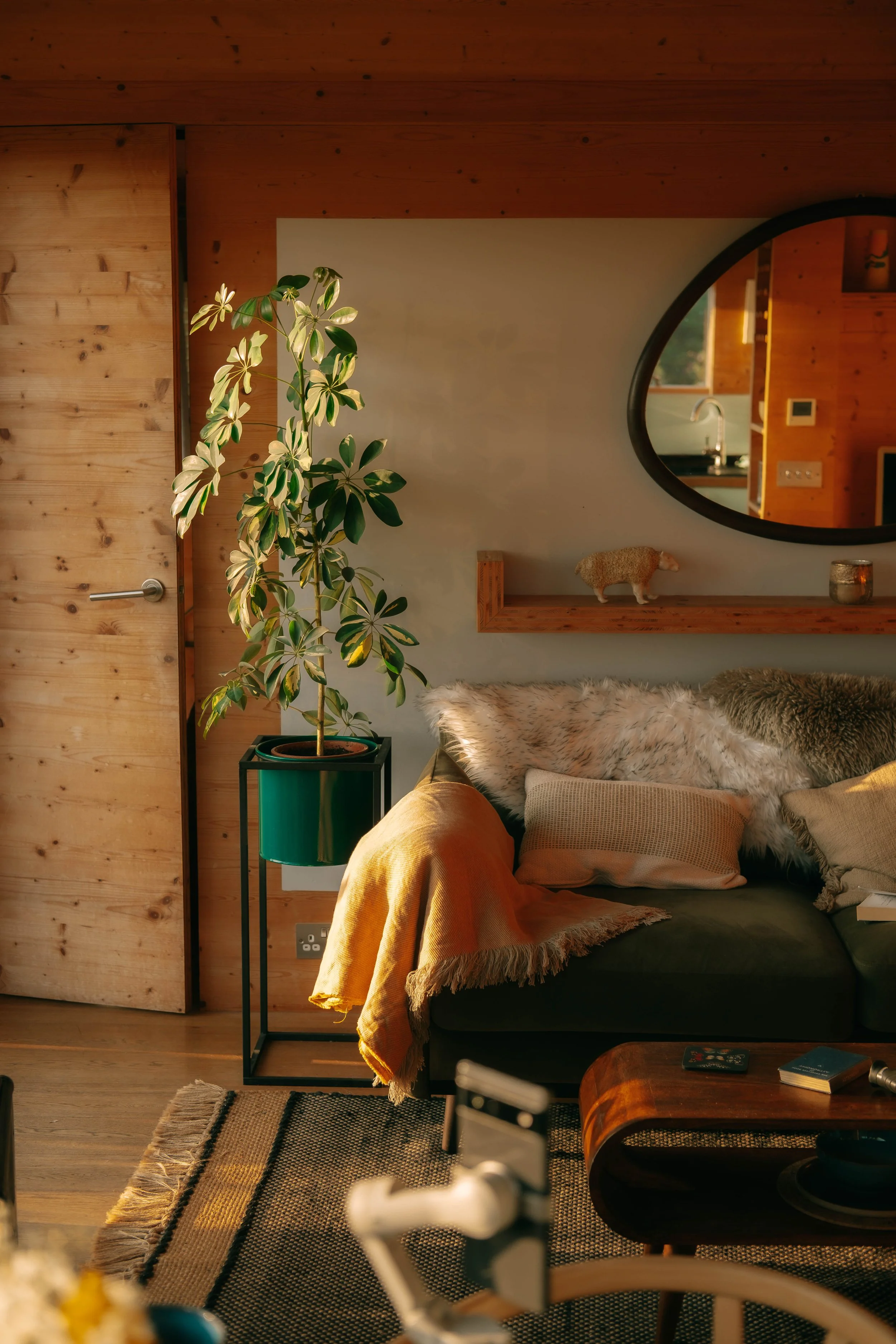 Living room with wooden walls, a green potted plant on a stand, a round mirror, a cozy sofa with fur and cushion, a wooden shelf with a small sheep figurine, a coffee table with books and remote, and a patterned rug.