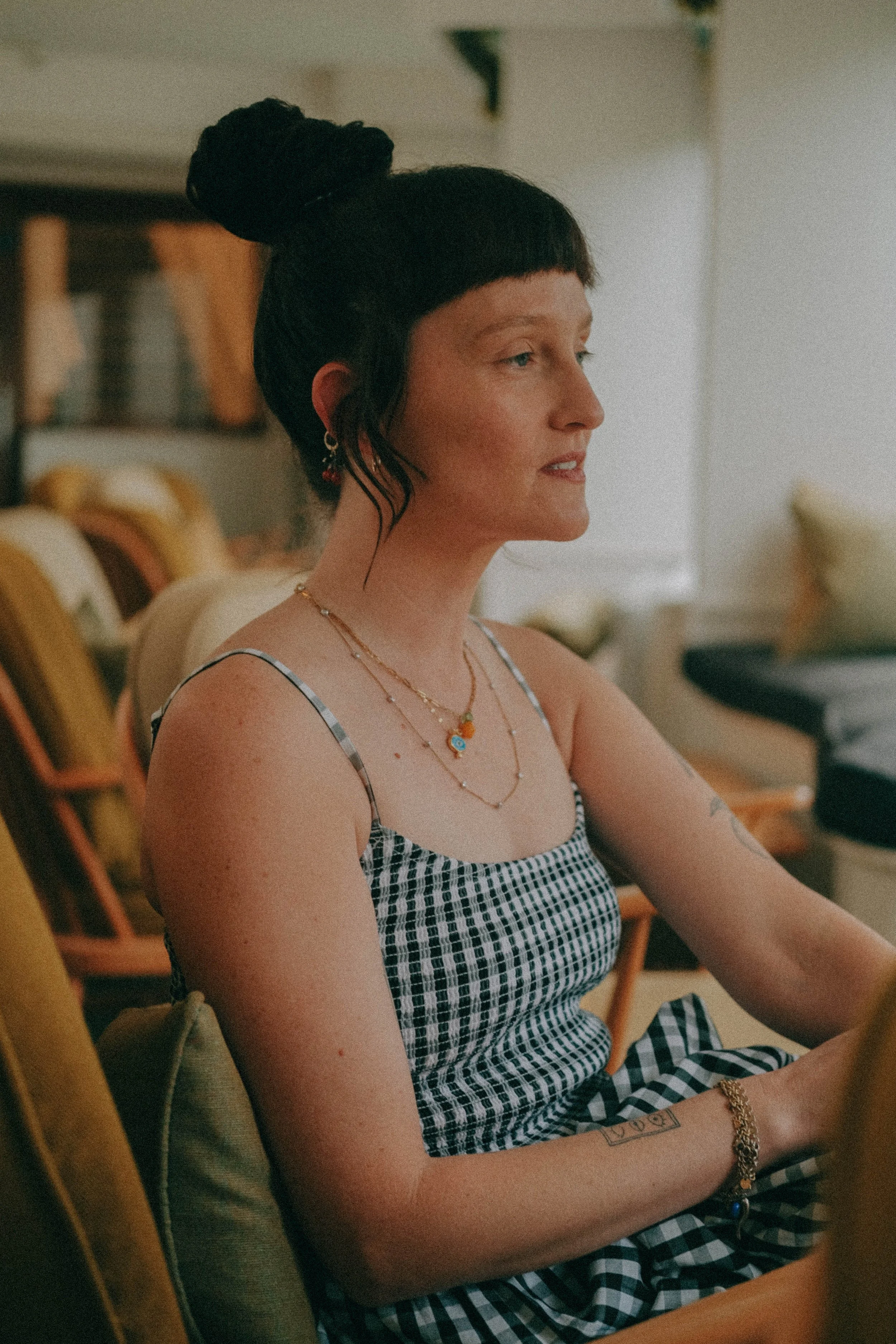 A woman with dark hair styled in a bun and bangs, wearing layered necklaces, earrings, a checkered tank top, and a bracelet, sitting indoors in a cozy, well-lit room with chairs and bookshelves in the background.