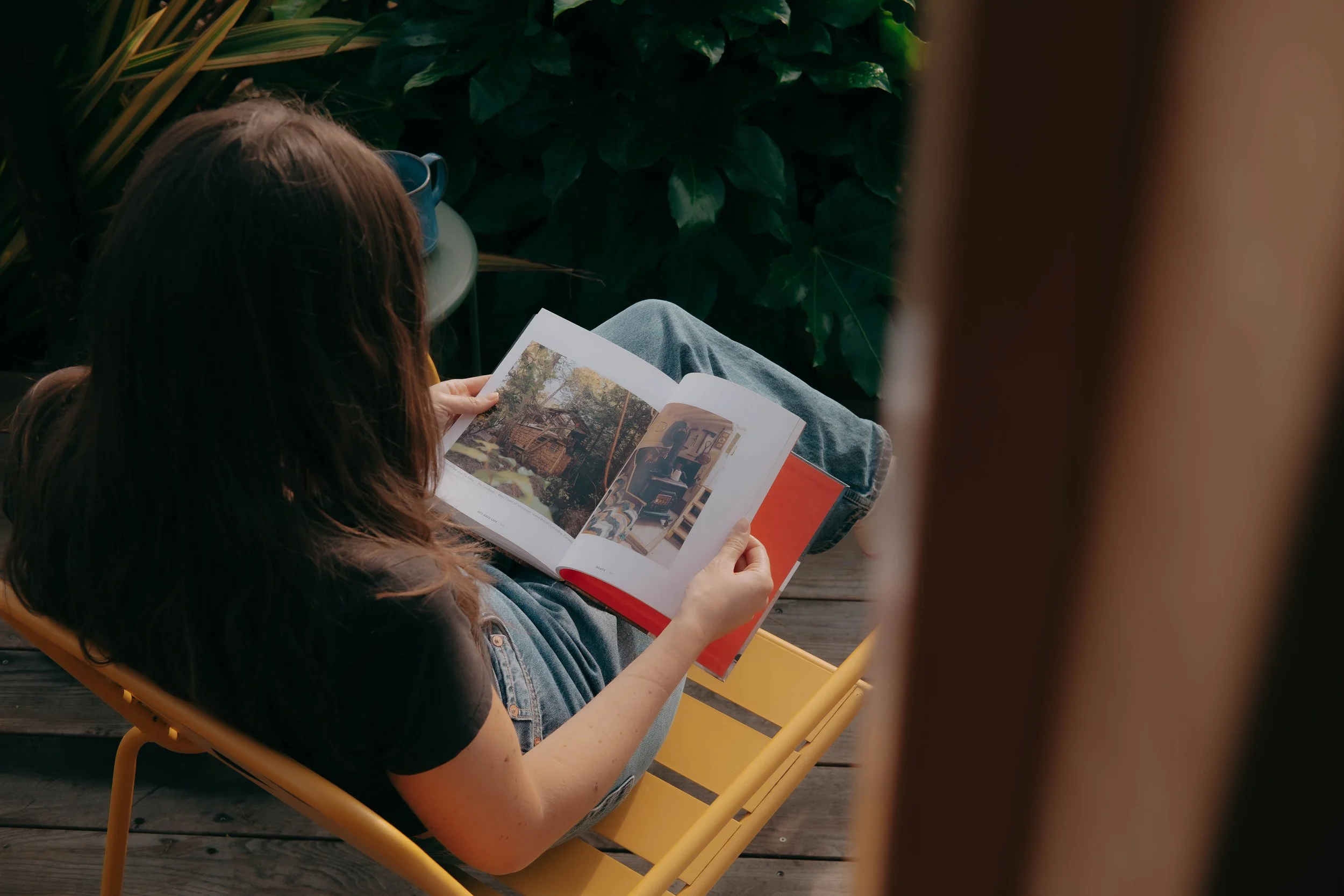 A woman sitting on a yellow chair outdoors, reading a picture magazine with several images of a house and an interior.