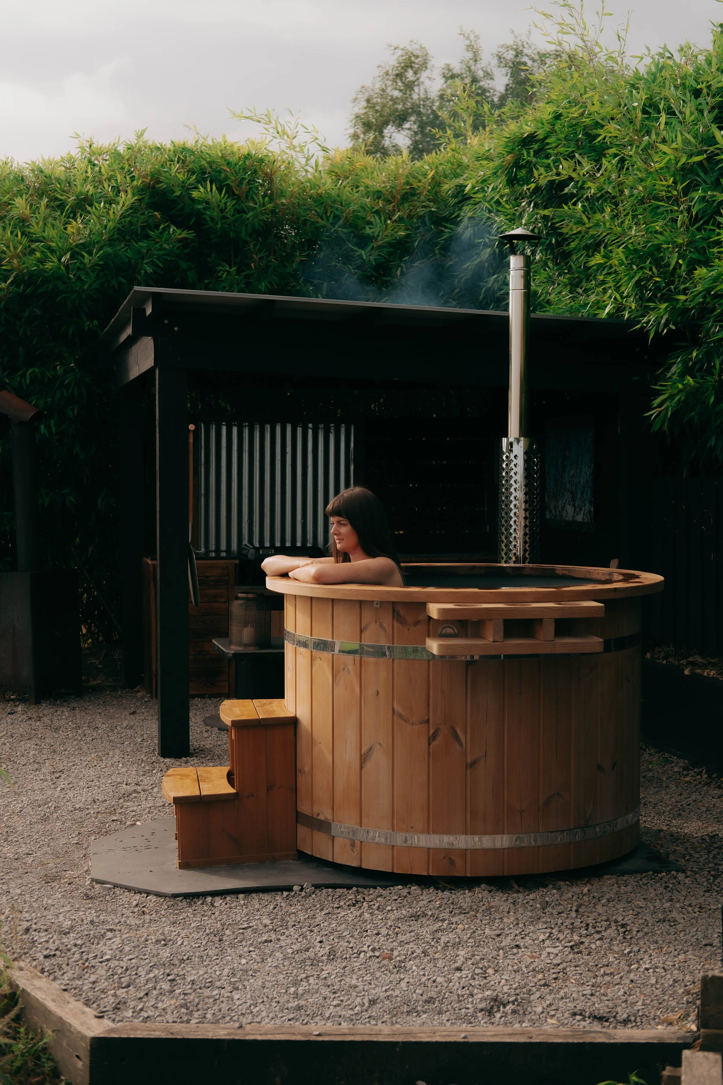 Woman relaxing in a wooden hot tub outdoors, surrounded by greenery.