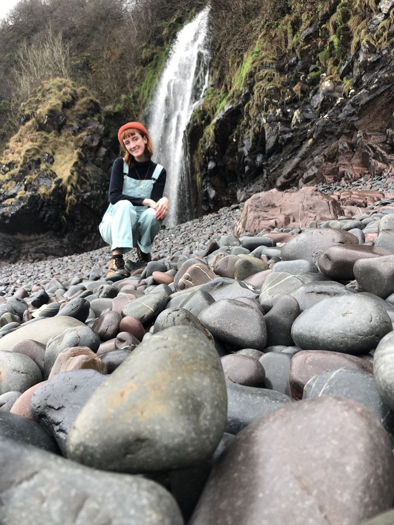 A smiling young woman wearing a peach hat and light blue overalls crouches on a rocky riverbed with a waterfall cascading down a mossy cliff behind her.