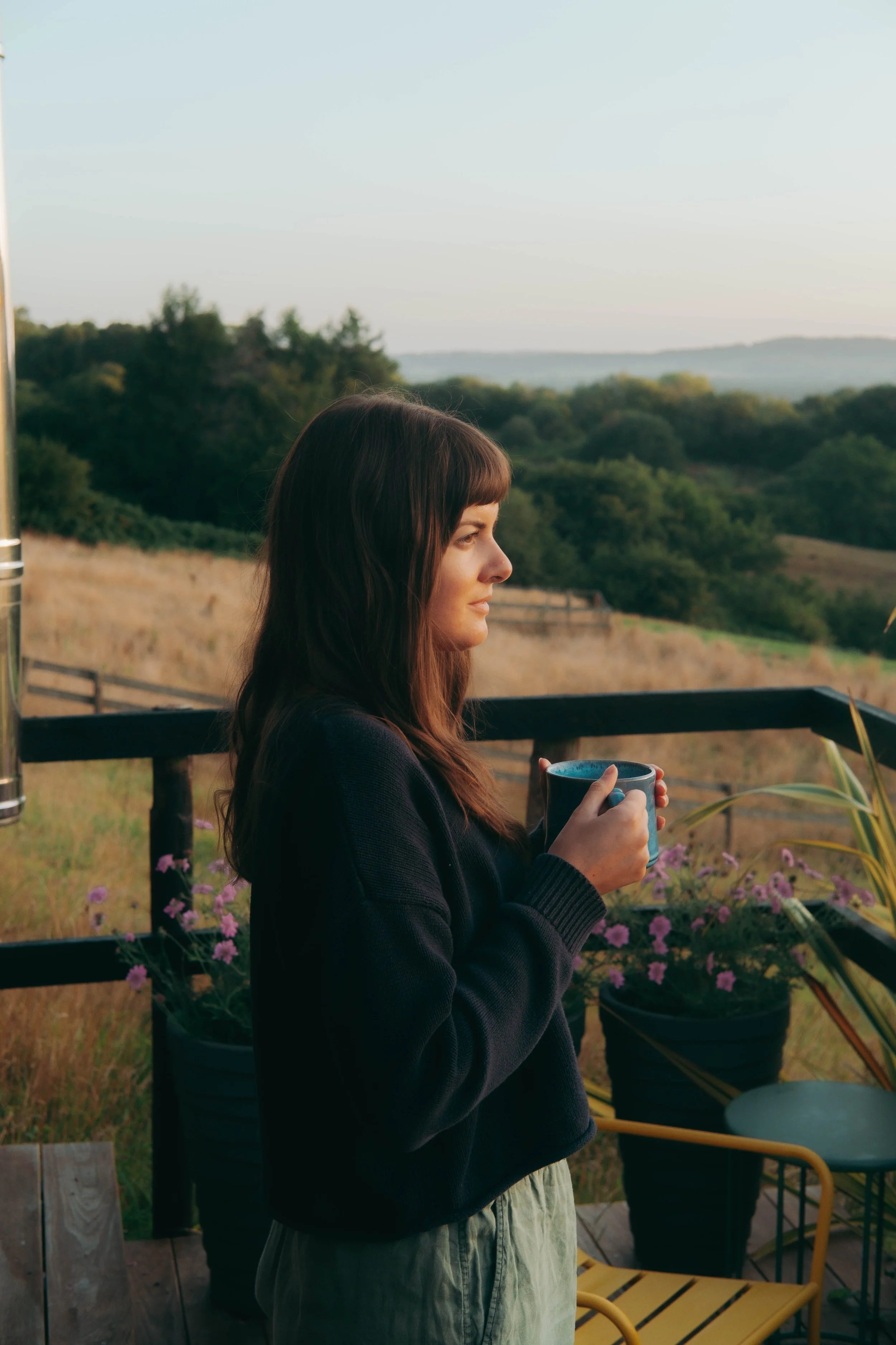 A woman with brown hair and bangs, wearing a dark sweater, stands on a wooden deck holding a blue mug, with rolling hills, trees, and a clear sky in the background.