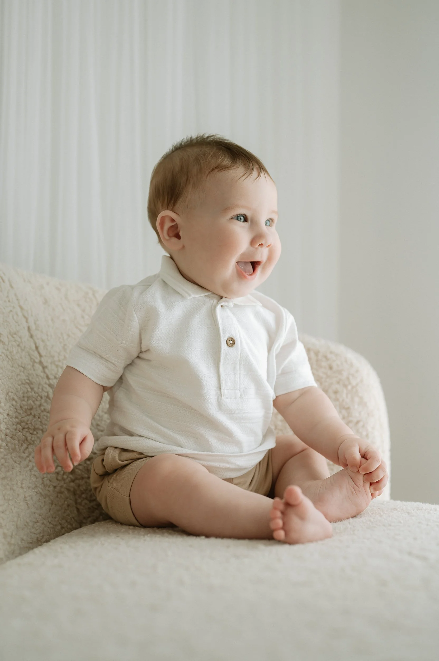 Smiling baby sitting on a cream sofa during a natural light sitter photography session at Alexis Stevenson Photography in Doncaster. Neutral, timeless baby portrait with soft tones, perfect for families looking for relaxed baby photography in South Y
