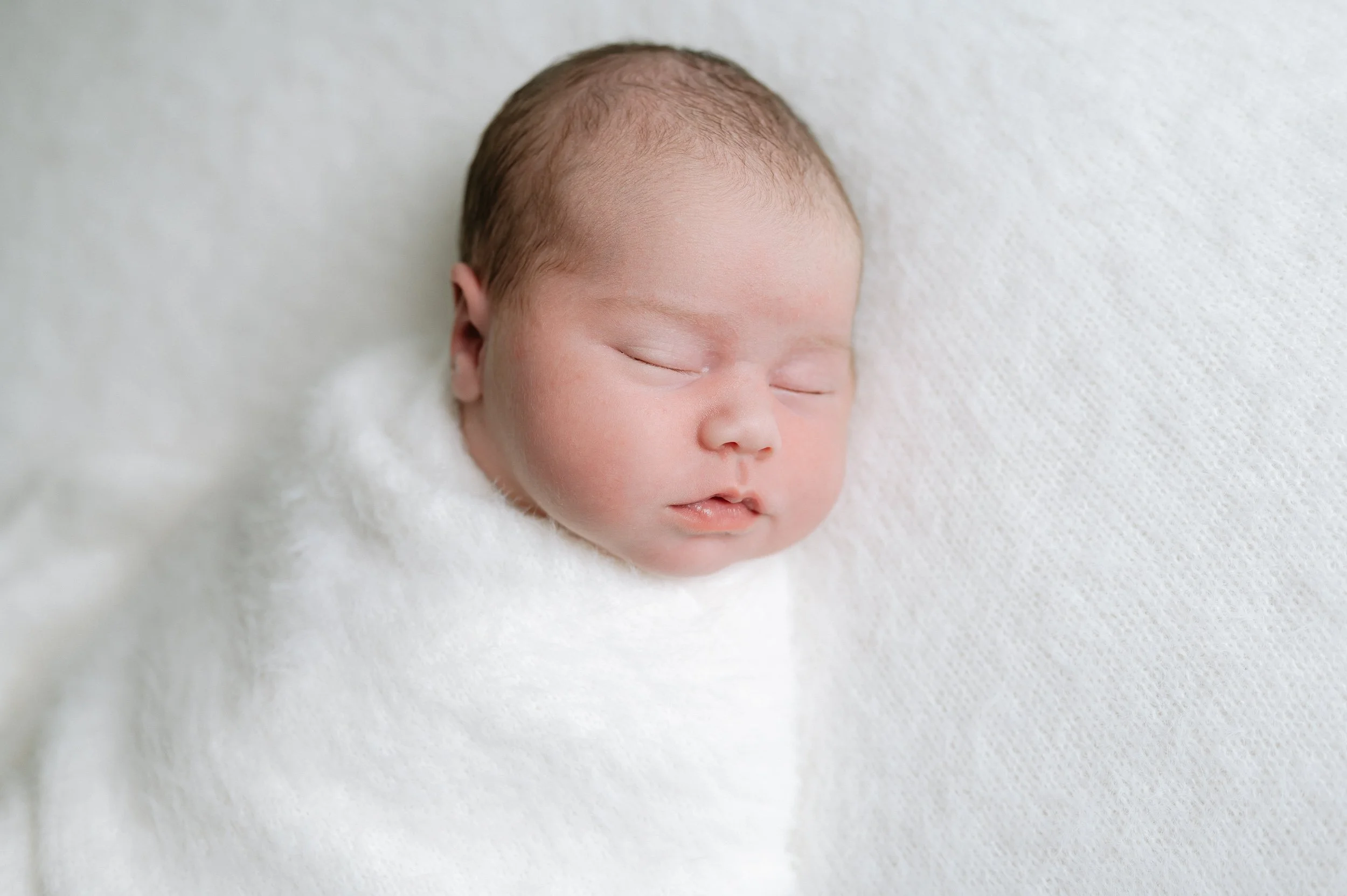 Portrait of a peacefully sleeping newborn baby wrapped in a soft white blanket during a natural light newborn photography session in Doncaster.