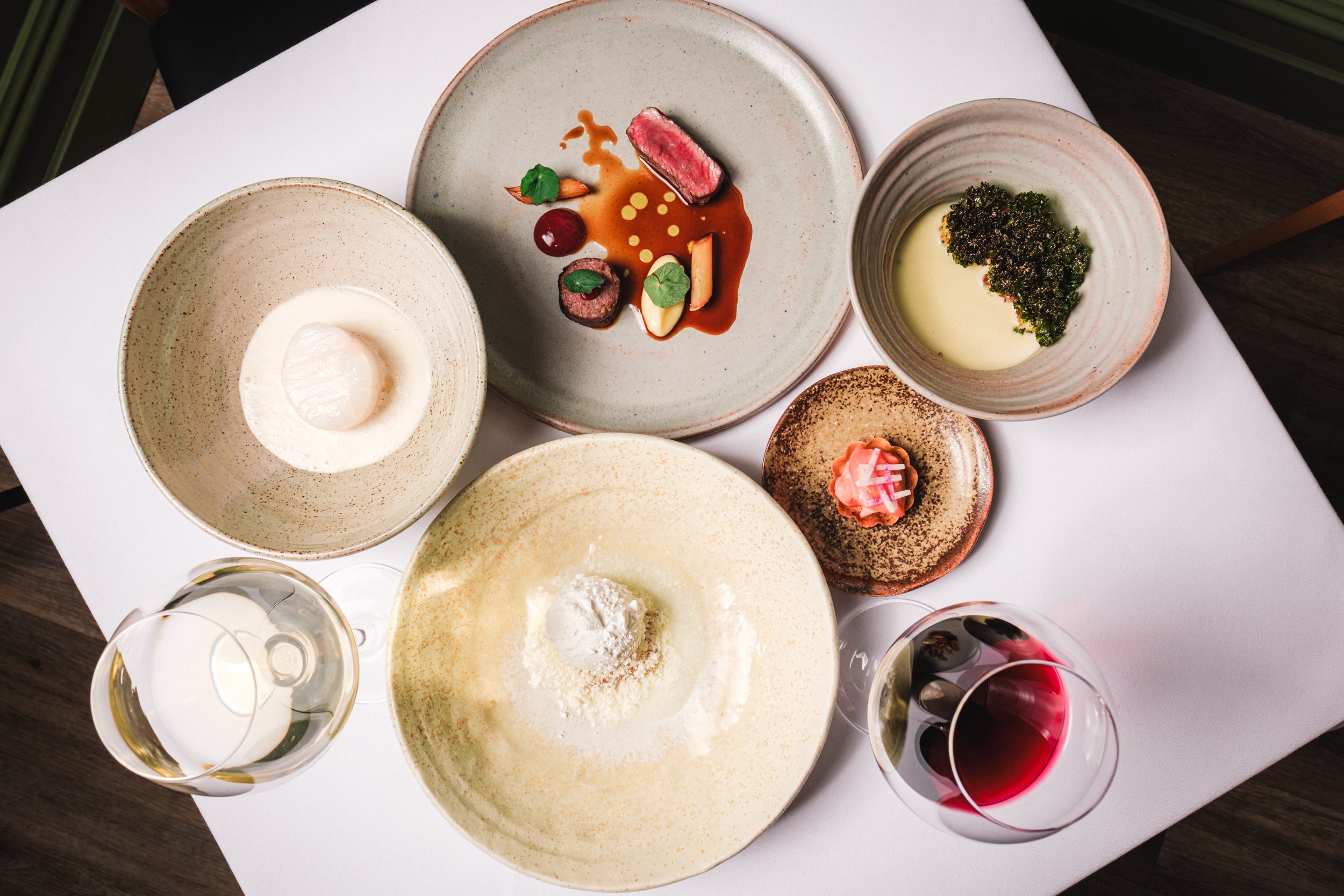 Gourmet meal with various dishes on plates and bowls, includes beef, scallop in sauce, leafy green garnish, small tart with radishes, and two wine glasses filled with red and white wine, on a white tablecloth.