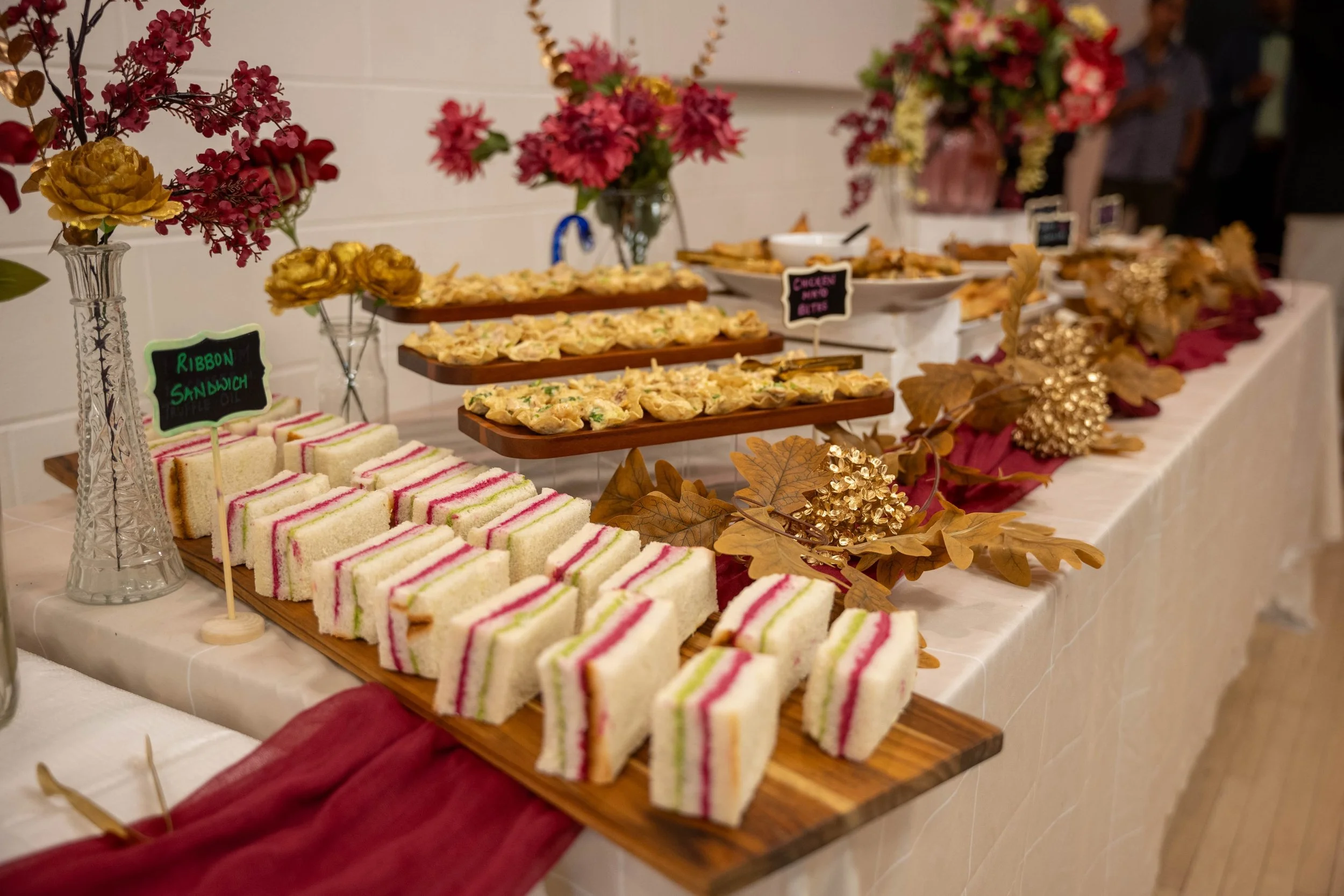 Assorted tacos on a table with small chalkboard signs labeled 'Chicken Xacuti', 'Chicken Cauliflower', and 'Navrti' at a party or event.