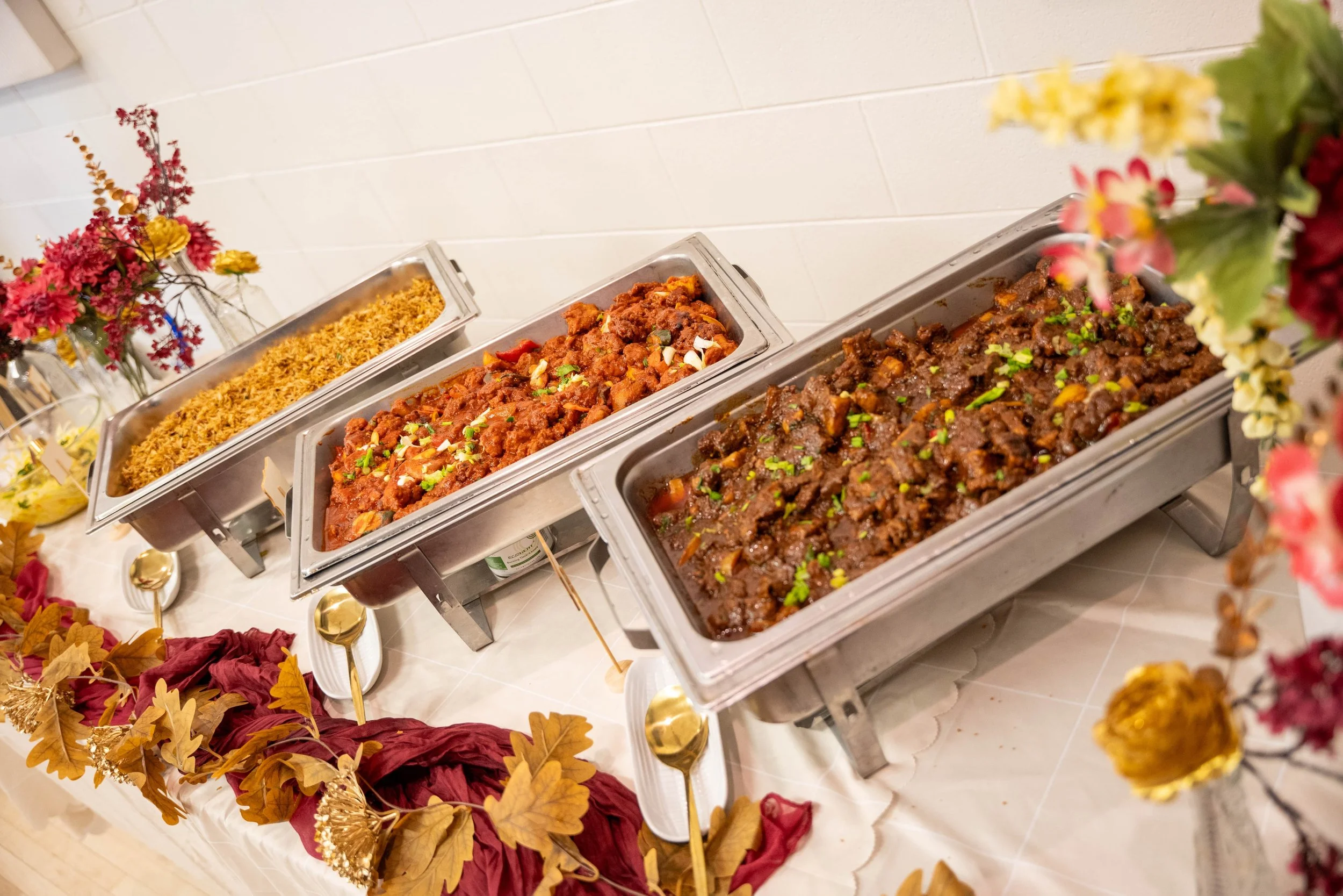 Buffet table with chafing dishes containing chicken, cauliflower, wings, and other foods, with lemon slices and small bowls of sauce.