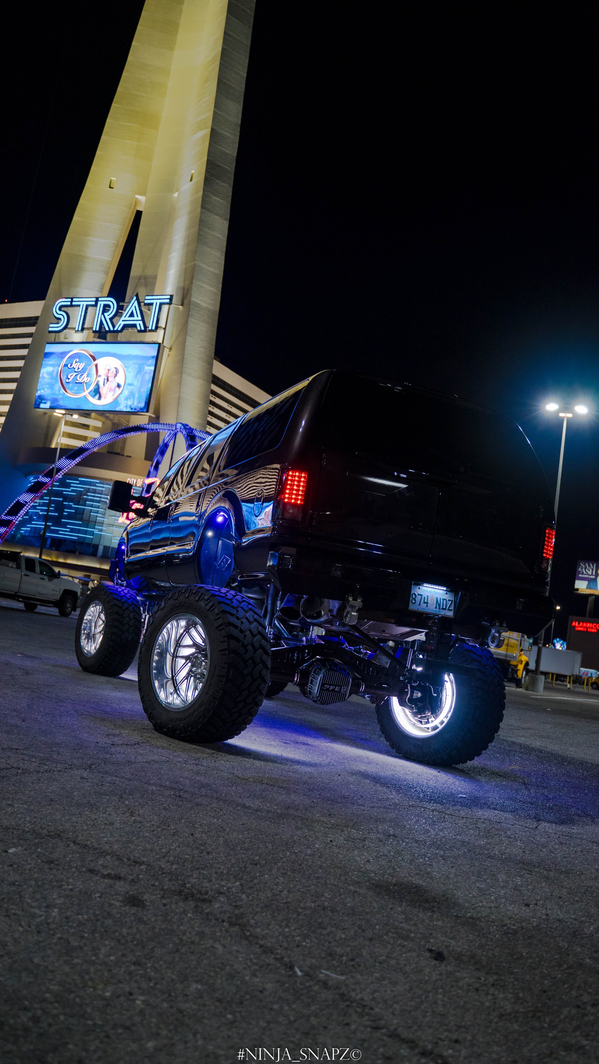A black SUV with oversized wheels lifted high off the ground parked in a lot at night, with bright city lights and signs in the background.