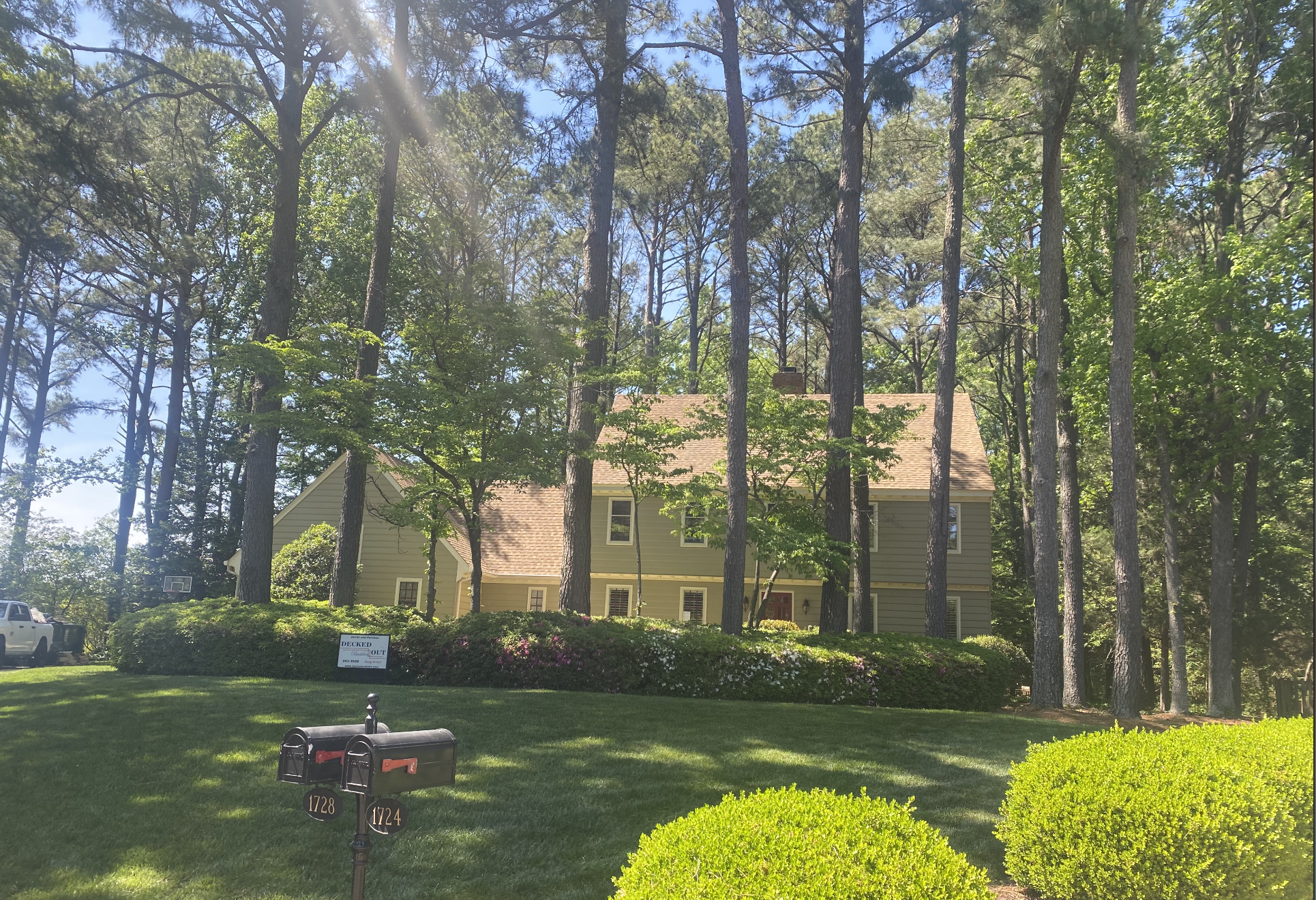 A beige house with a pitched roof is surrounded by tall trees and well-manicured bushes, with a bright sky overhead. There are two black mailboxes with house numbers 1728 and 1724 in the foreground, and a small sign that says 'Decked Out' near the bu