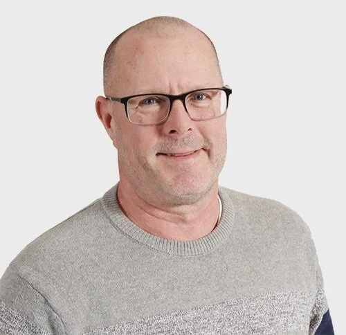 Portrait of a middle-aged man with glasses, short hair, wearing a gray sweater, smiling against a white background.