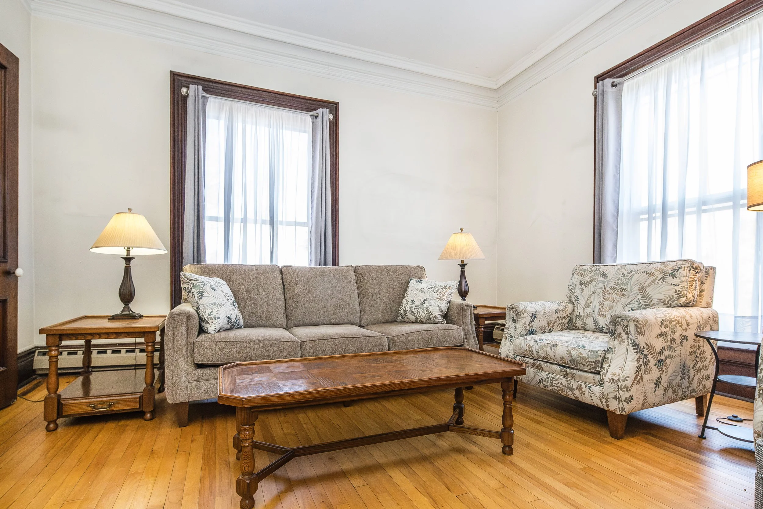 Living room with beige sofa, patterned armchair, wooden coffee table, side tables, lamps, and windows with white curtains.