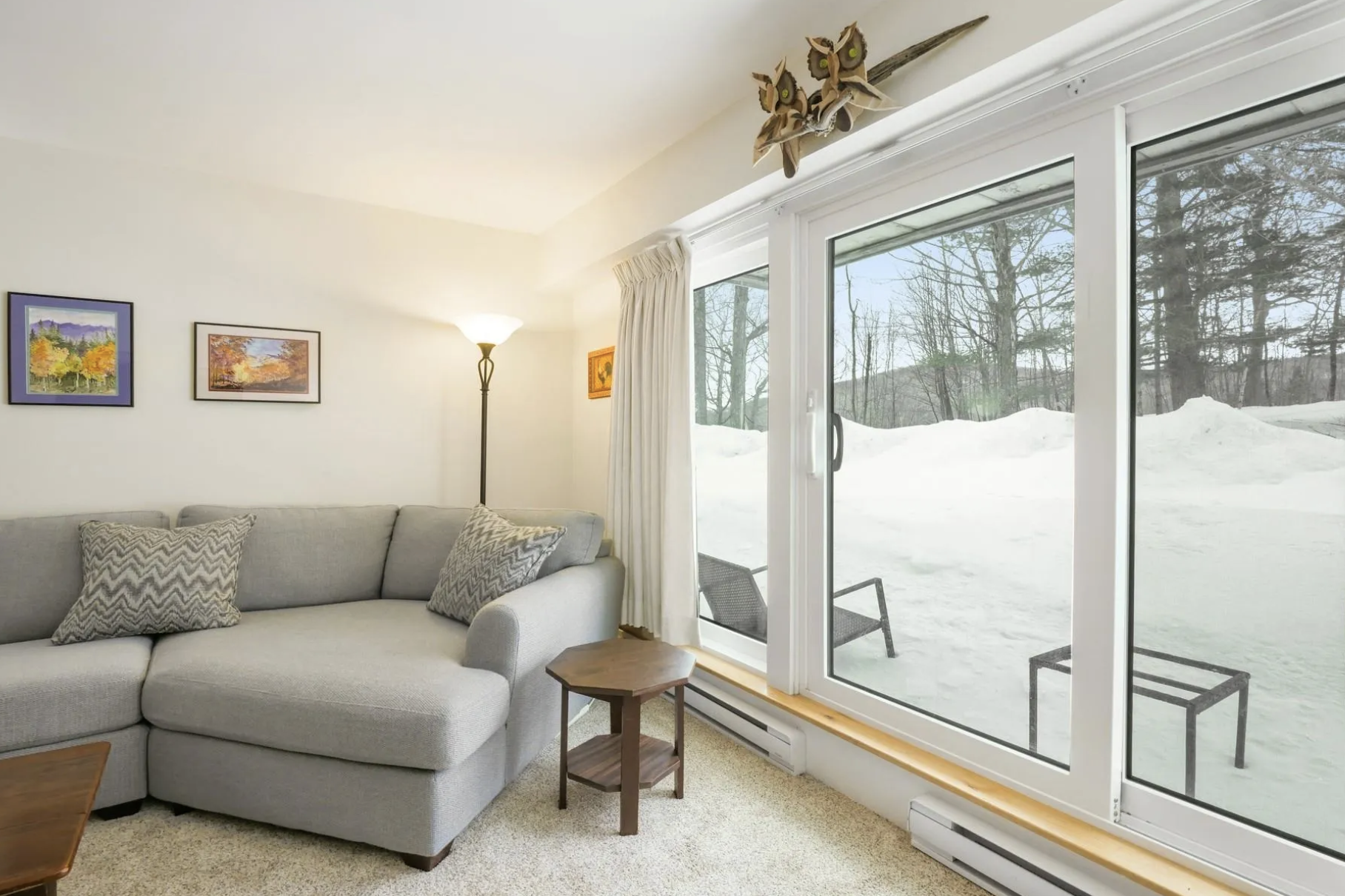 Living room with gray sectional sofa, patterned pillows, a small wooden side table, a tall floor lamp, wall art, and a large window showing snow outside with two chairs on the patio.