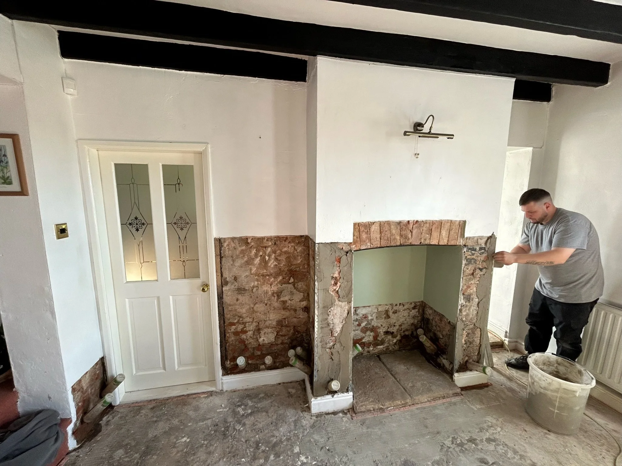 A man working on a home renovation project, removing bricks from a fireplace. The room has a white door with glass panels, white walls, black ceiling beams, and a bucket of construction materials on the floor.