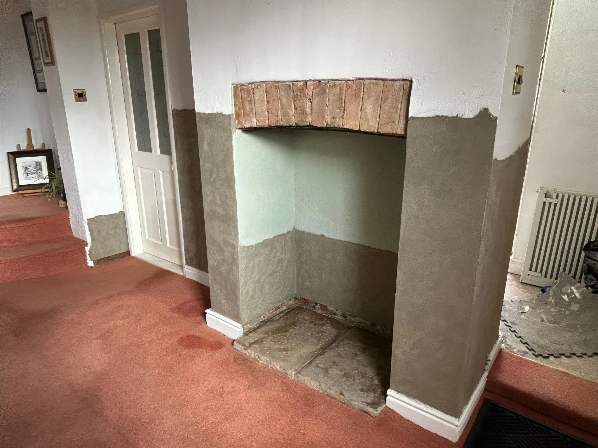 Interior of a house under renovation showing a partially built fireplace with a brick arch, plastered sides, and a concrete hearth. The carpeted floor is reddish-orange and some furniture and framed pictures are visible in the background.
