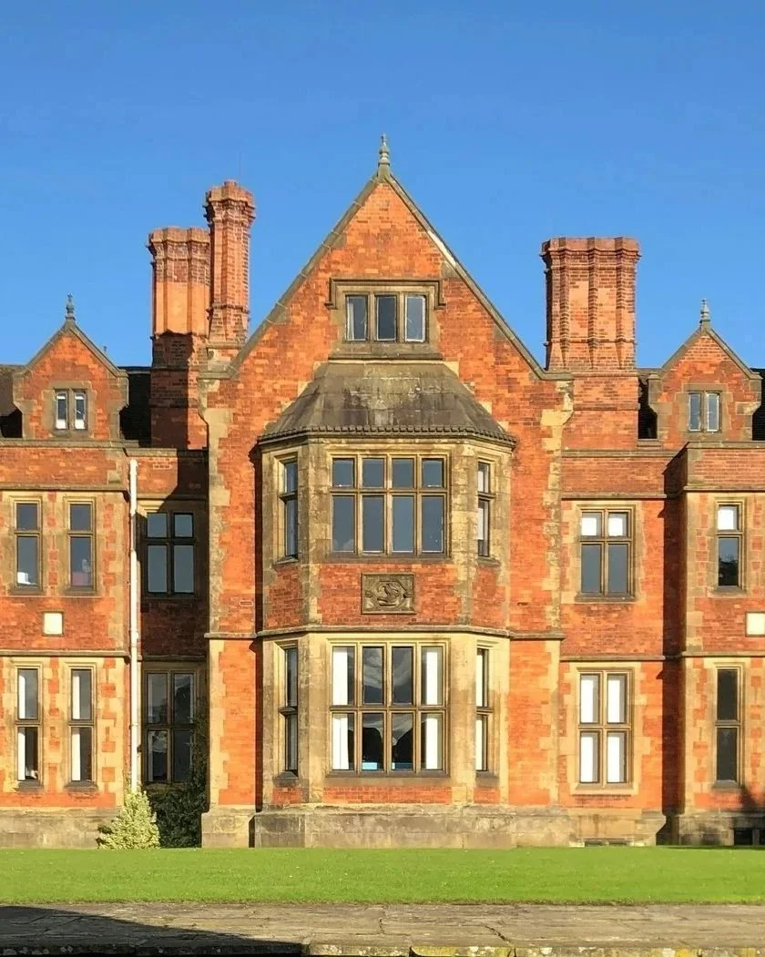 A historic red brick building with ornate window frames, multiple chimneys, and a decorative stone crest, set against a clear blue sky and green lawn.