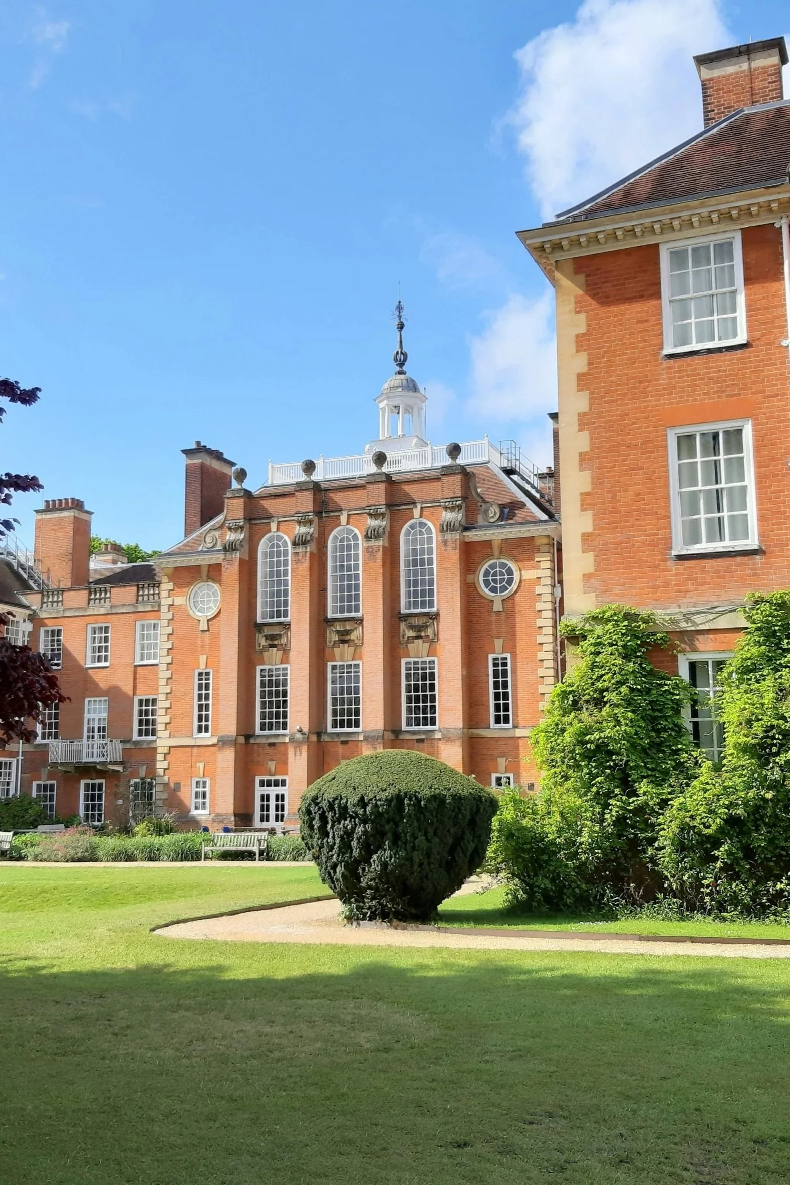 A large brick building with multiple windows and a decorative turret on top, set against a clear blue sky, with a well-maintained garden featuring neatly trimmed bushes and green grass in the foreground.