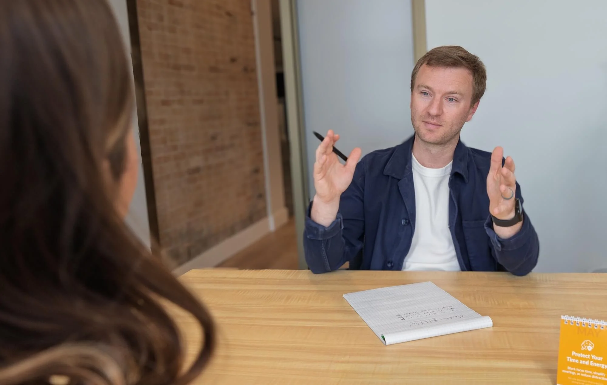 A man with light brown hair and a beard sitting at a table, engaging in conversation with a woman with brown hair, seen from behind. The man is holding a black pen, wearing a blue jacket over a white shirt, and sitting in a modern office or meeting room with a brick wall and whiteboard in the background. There are notebooks and a calendar on the table.
