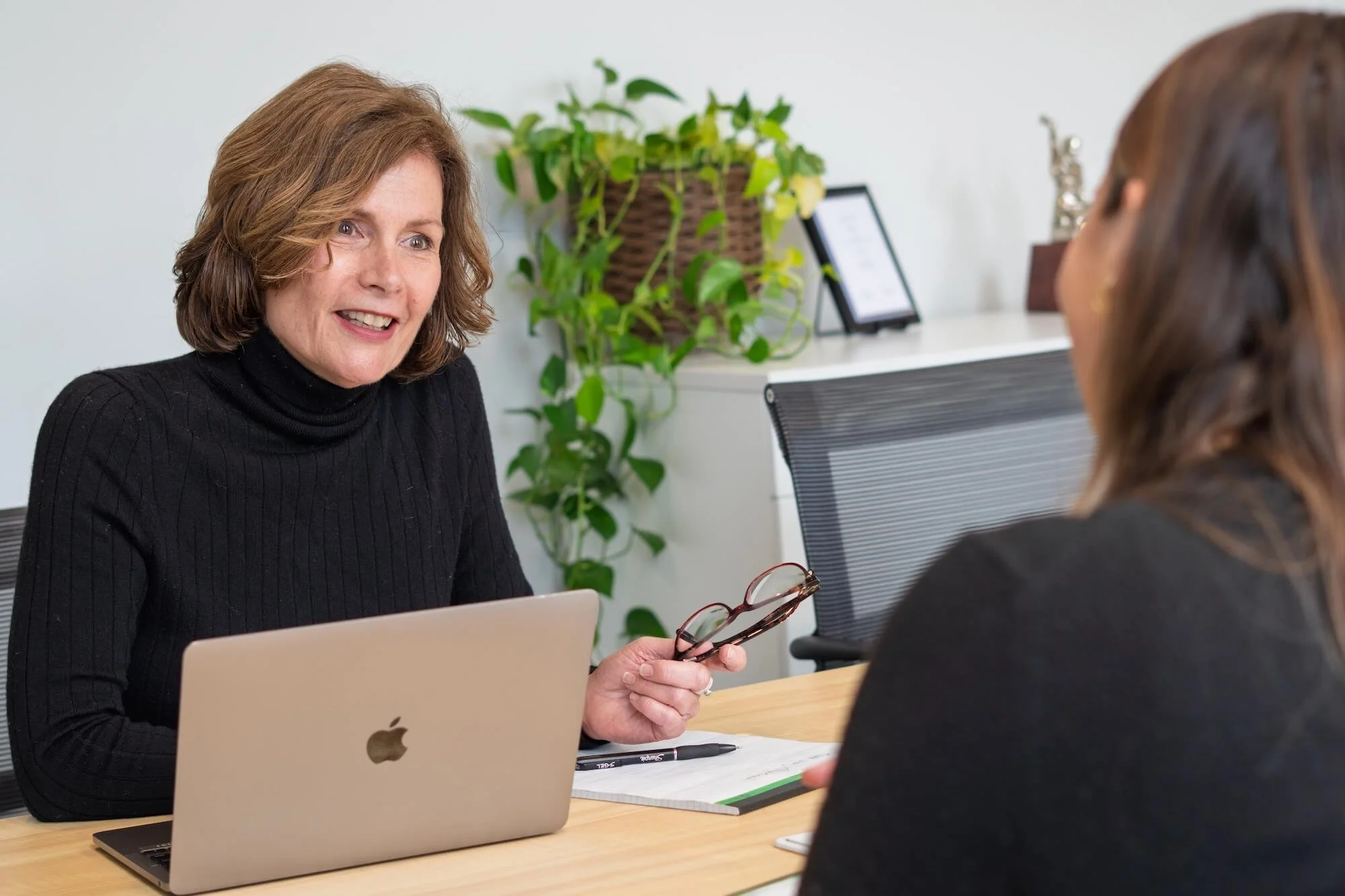 Two women are engaged in a conversation in an office setting. One woman, with shoulder-length brown hair, is smiling and holding glasses, while the other woman, with long brown hair, is seen from behind. There is a laptop, a notepad, pens, and a potted plant on the desk, with a framed picture and decorative items on the white wall in the background.
